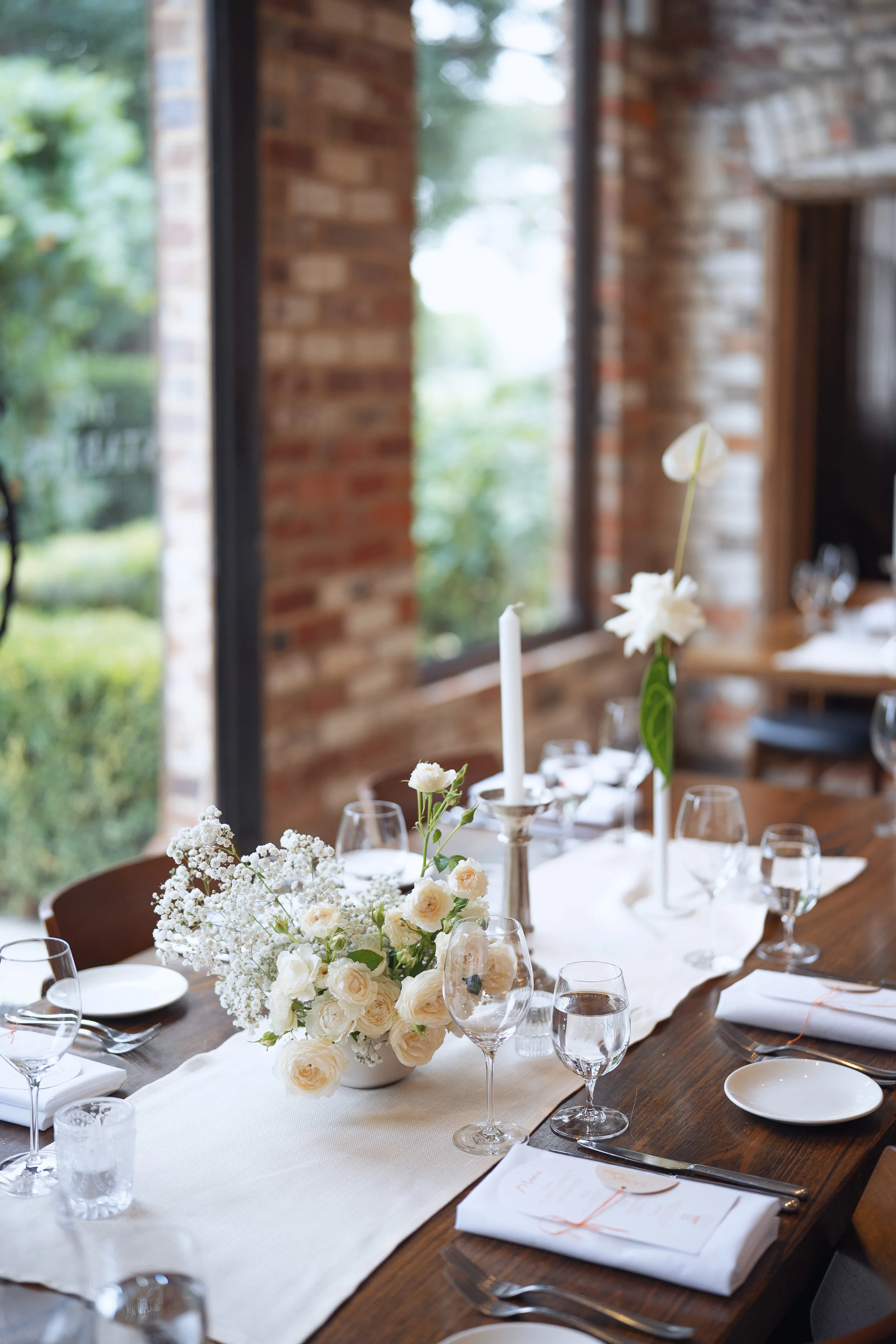 A decorated dining table with a floral centerpiece, gold candle holders, wine glasses, plates, and napkins, set in a room with large windows and brick walls.