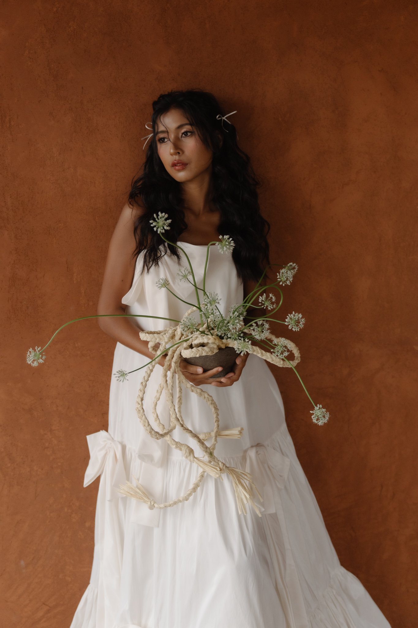 A woman in a white dress with black, wavy hair, holding a flower arrangement with white flowers and long green stems, standing against a brown wall.