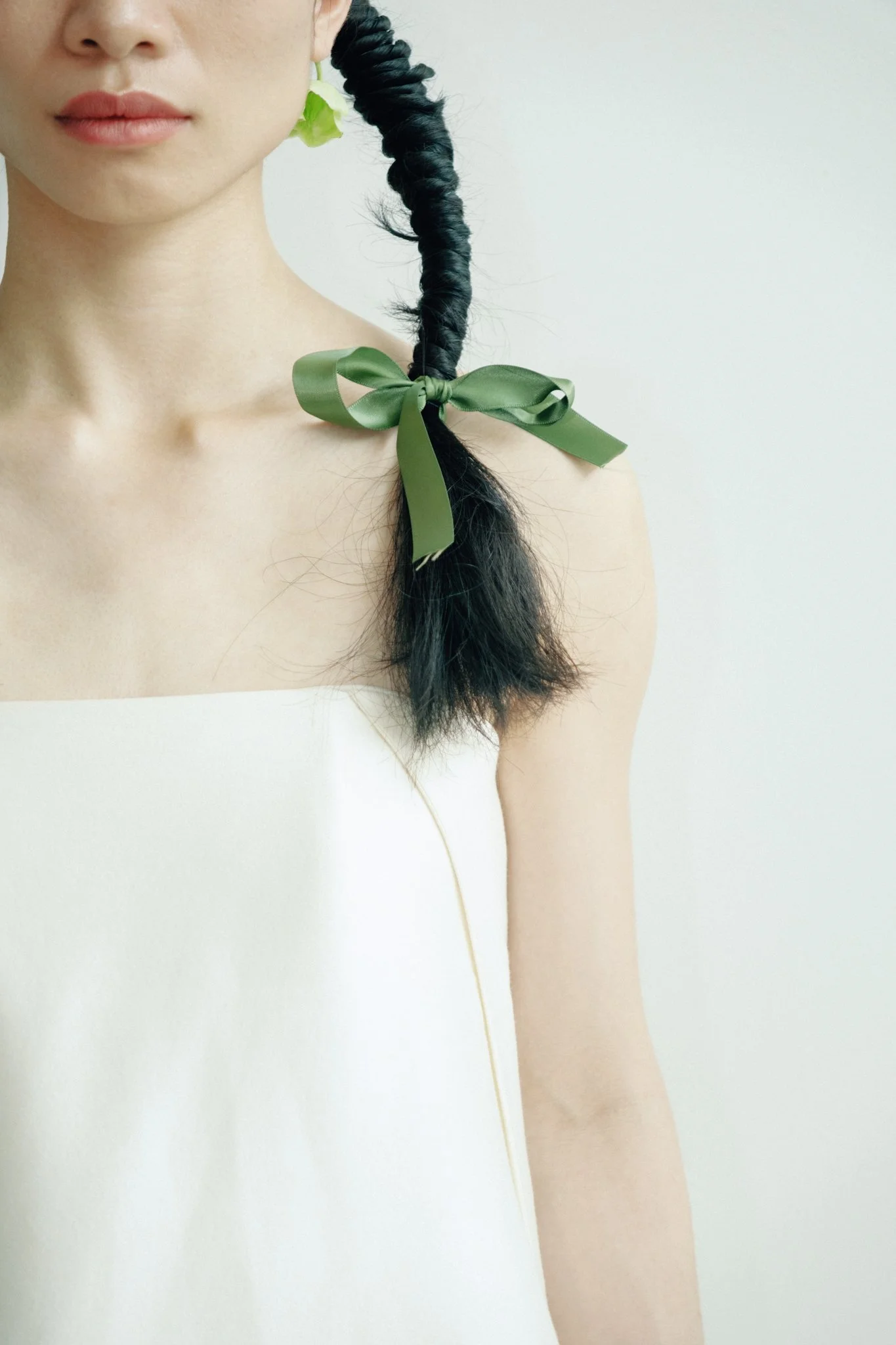 Close-up of a woman with long dark hair styled in a twisted side braid, tied with a green satin ribbon bow, wearing a white strapless top, with a plain light-colored background.