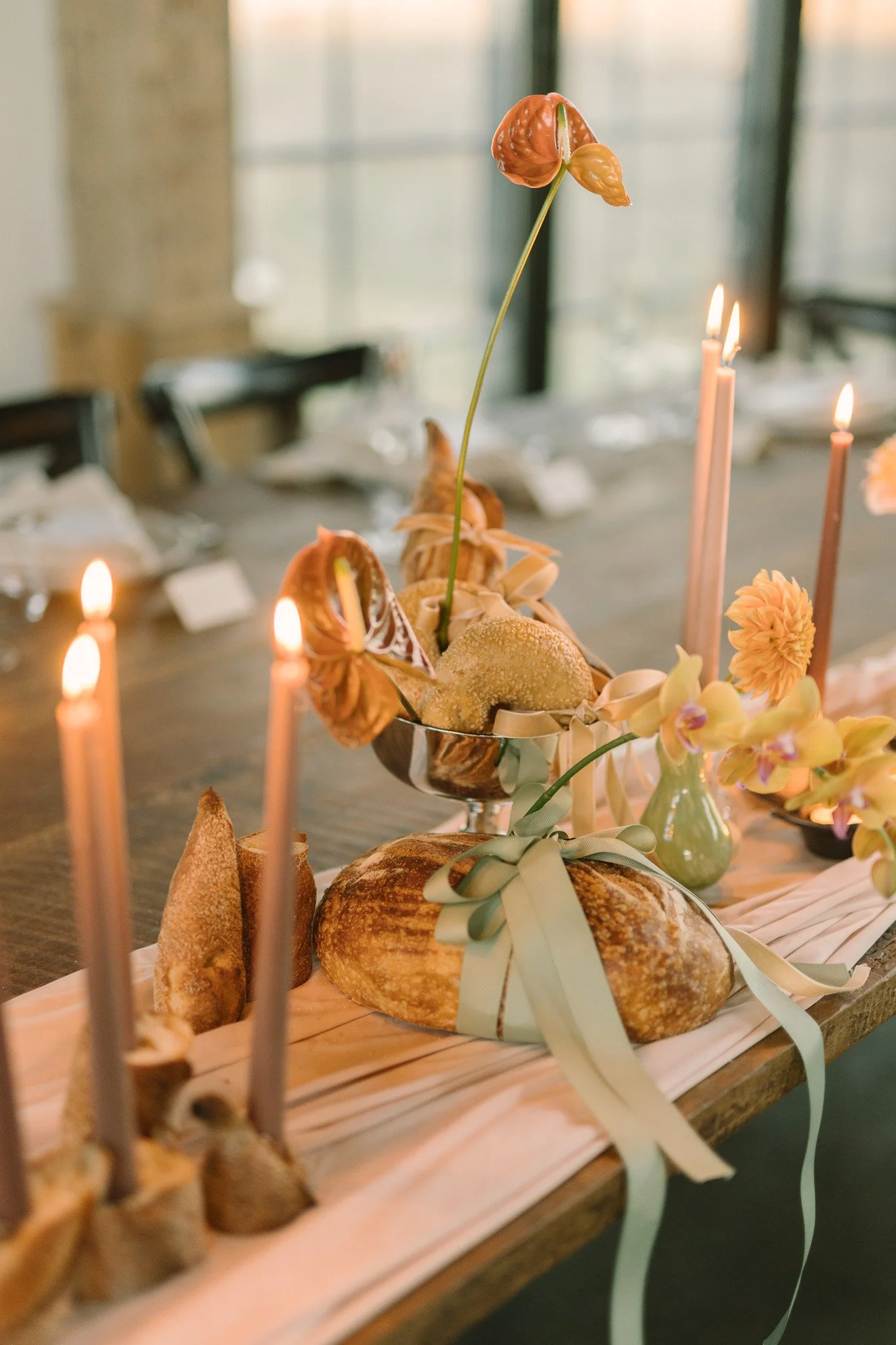 A table decorated with various breads, flowers, and tall pink candles, set in a bright room with large windows.