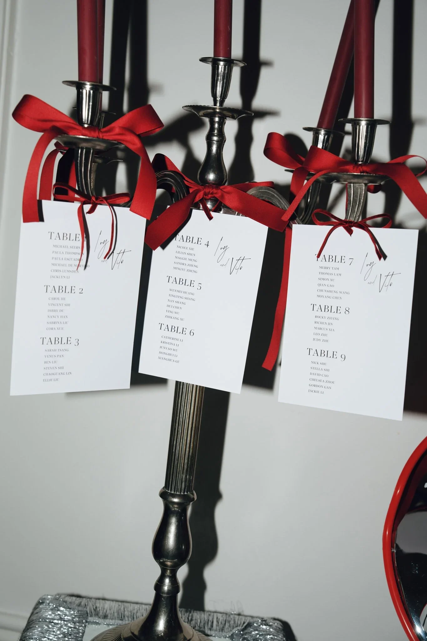 A metal candle holder with four red candles and red ribbons tied around its forks, holding seating cards with lists of names for a formal event. The candles cast shadows on the wall.