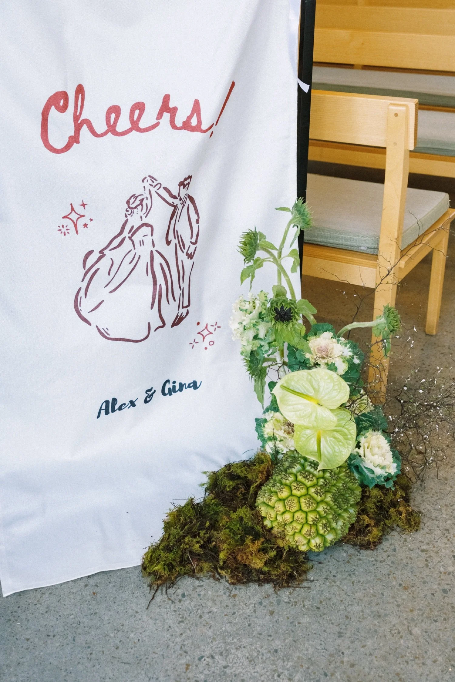 A floral arrangement with various green plants and flowers in front of a white cloth banner with the words 'Cheers!' and a drawing of a dancing couple, with wooden seating in the background.