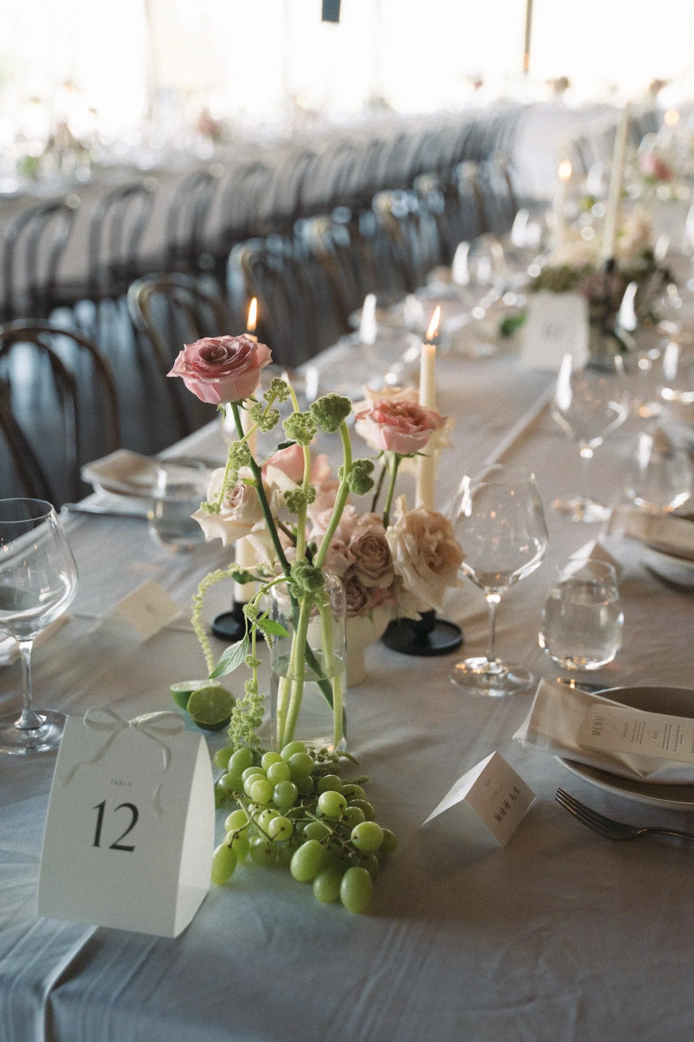 A floral centerpiece with pink roses, green berries, and greenery on a table at an event, surrounded by wine glasses, place settings, and table number 12.