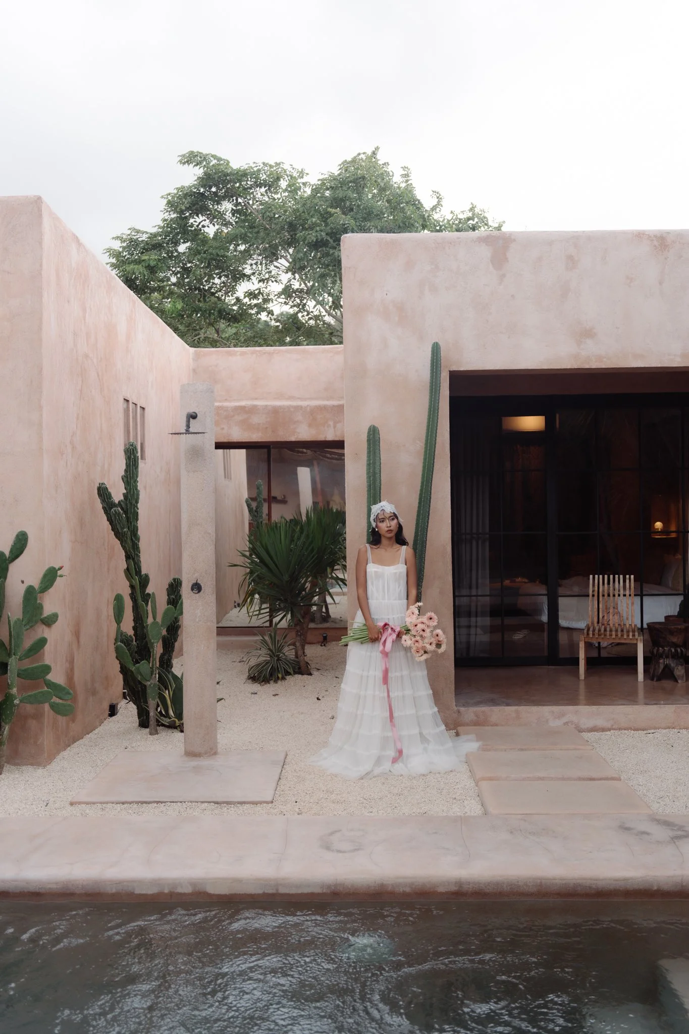 A woman in a white dress holding a bouquet of pink flowers, standing outside near a modern beige building with cacti and desert plants, beside a canal or waterway.