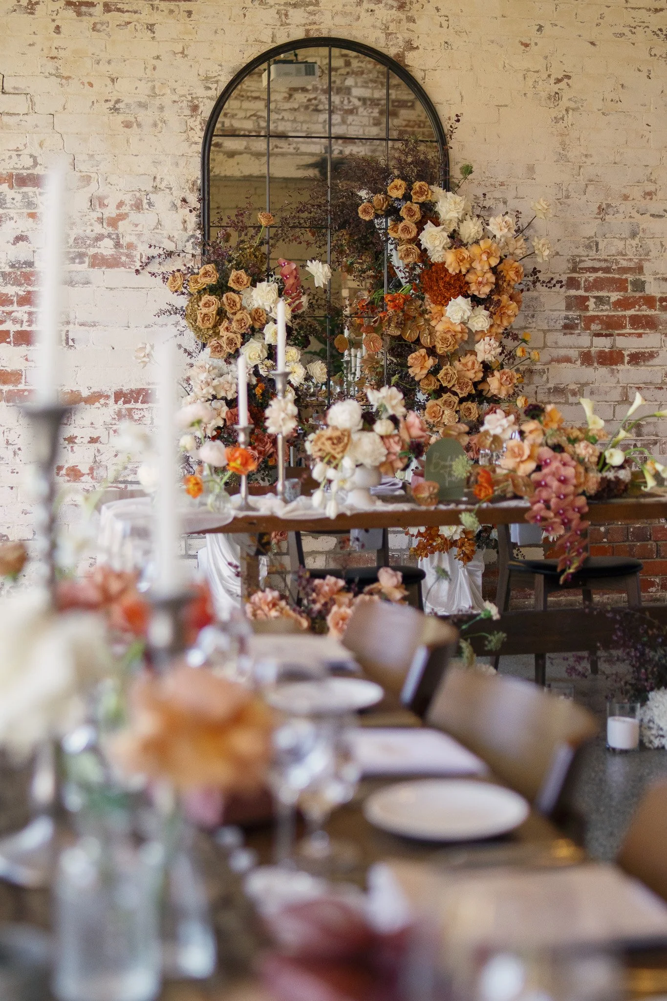 A decorated dining table with floral arrangements and candles in front of a brick wall and a large arched mirror.