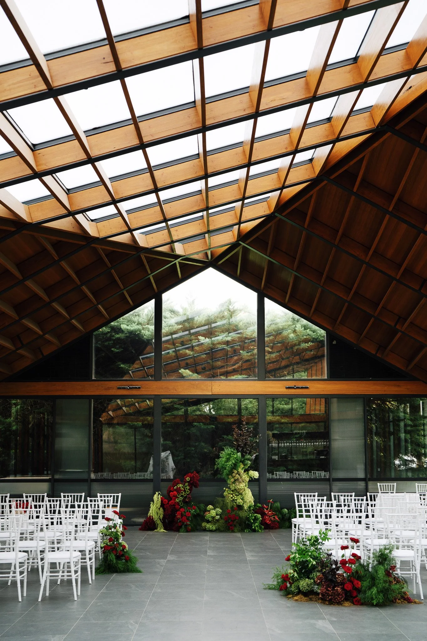 A modern indoor space with a high wooden ceiling that has skylights, and large glass windows showing greenery outside. The room is set up with rows of white chairs and decorated with colorful flower arrangements, suggesting it is prepared for a weddi