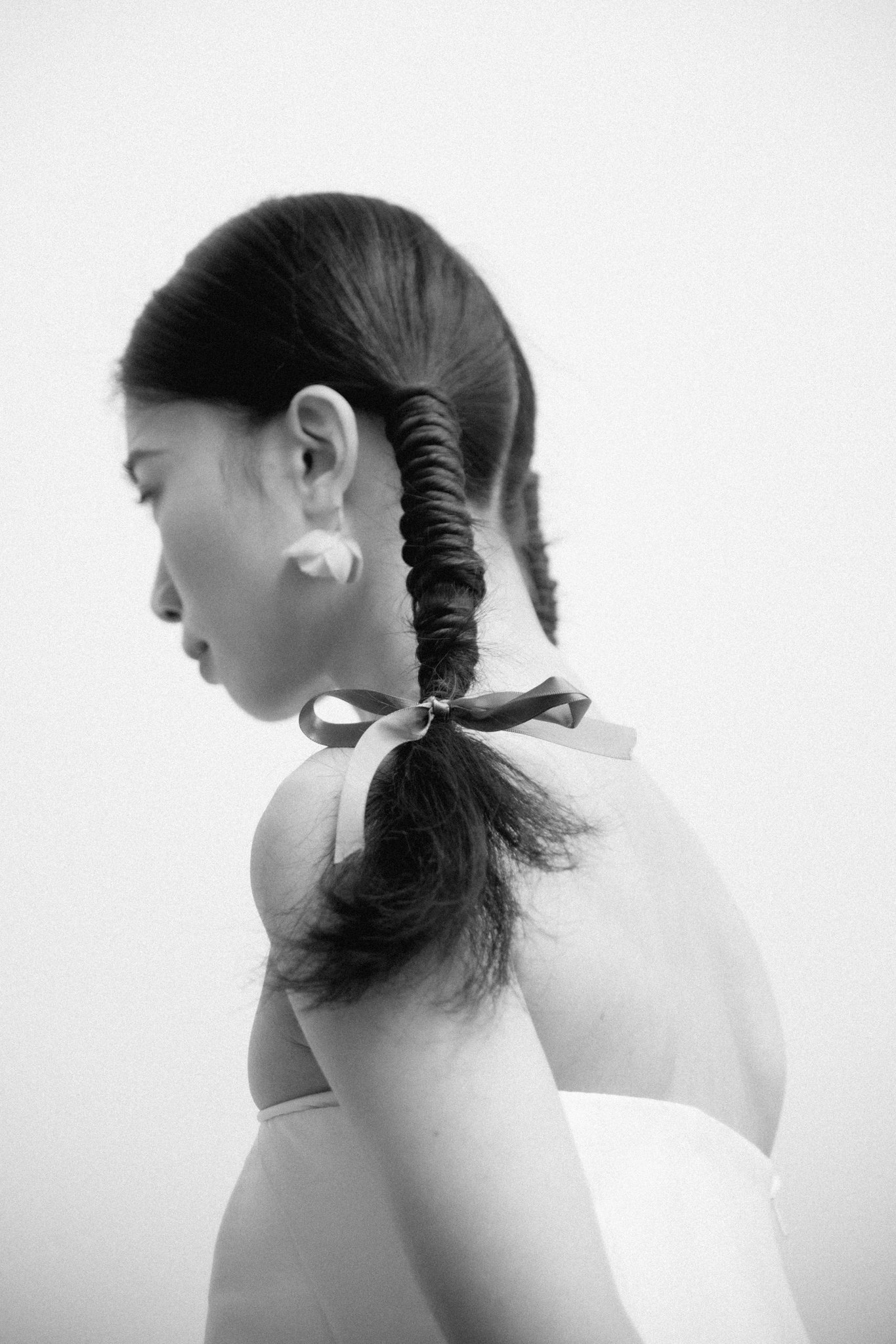 A black and white photograph of a woman with long hair styled in two twisted braids tied with ribbons, her eyes closed, against a plain light background.