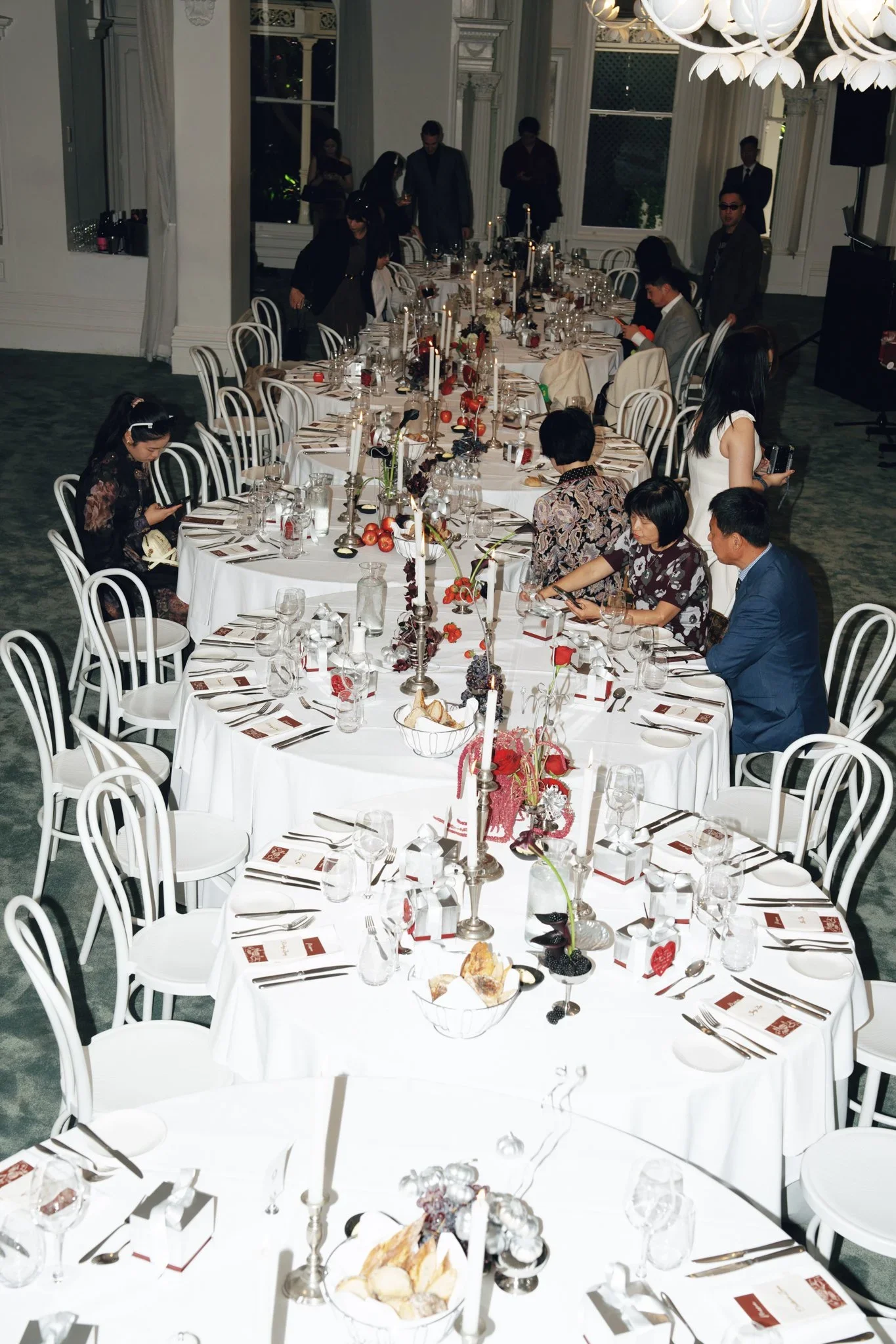 A long banquet table set for a meal with white tablecloths, candles, flowers, glassware, and cutlery. There are several people seated and some standing near the table in a large, elegant room with ornate white walls and large windows. The scene appea
