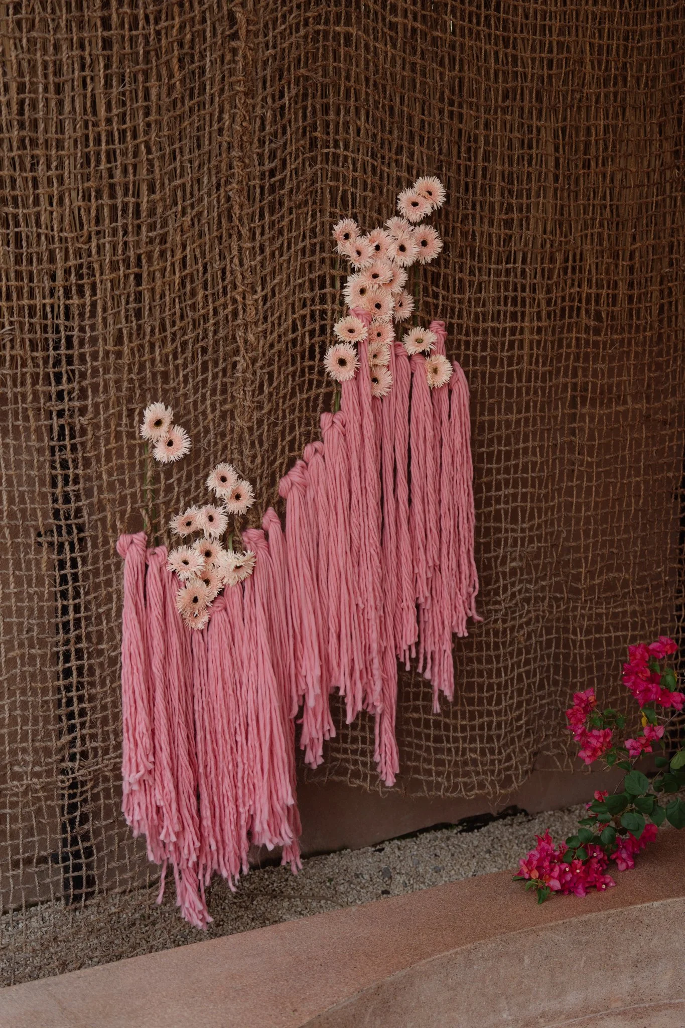 Pink artificial flowers with long pink yarns hanging down, set against a brown woven backdrop with pink flowering plants nearby.