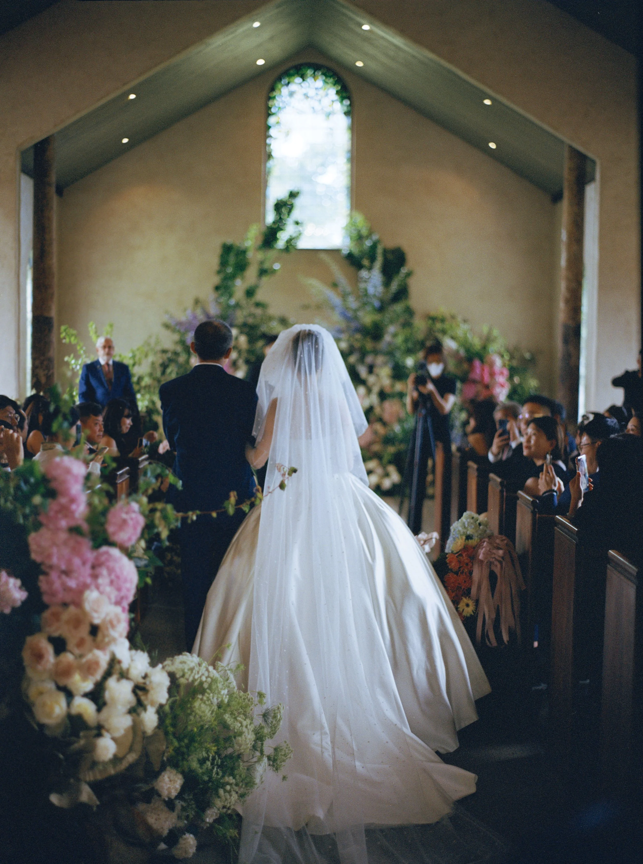 A couple getting married in a church, with a large floral arrangement and floral decorations along the aisle, and a stained glass window in the background.