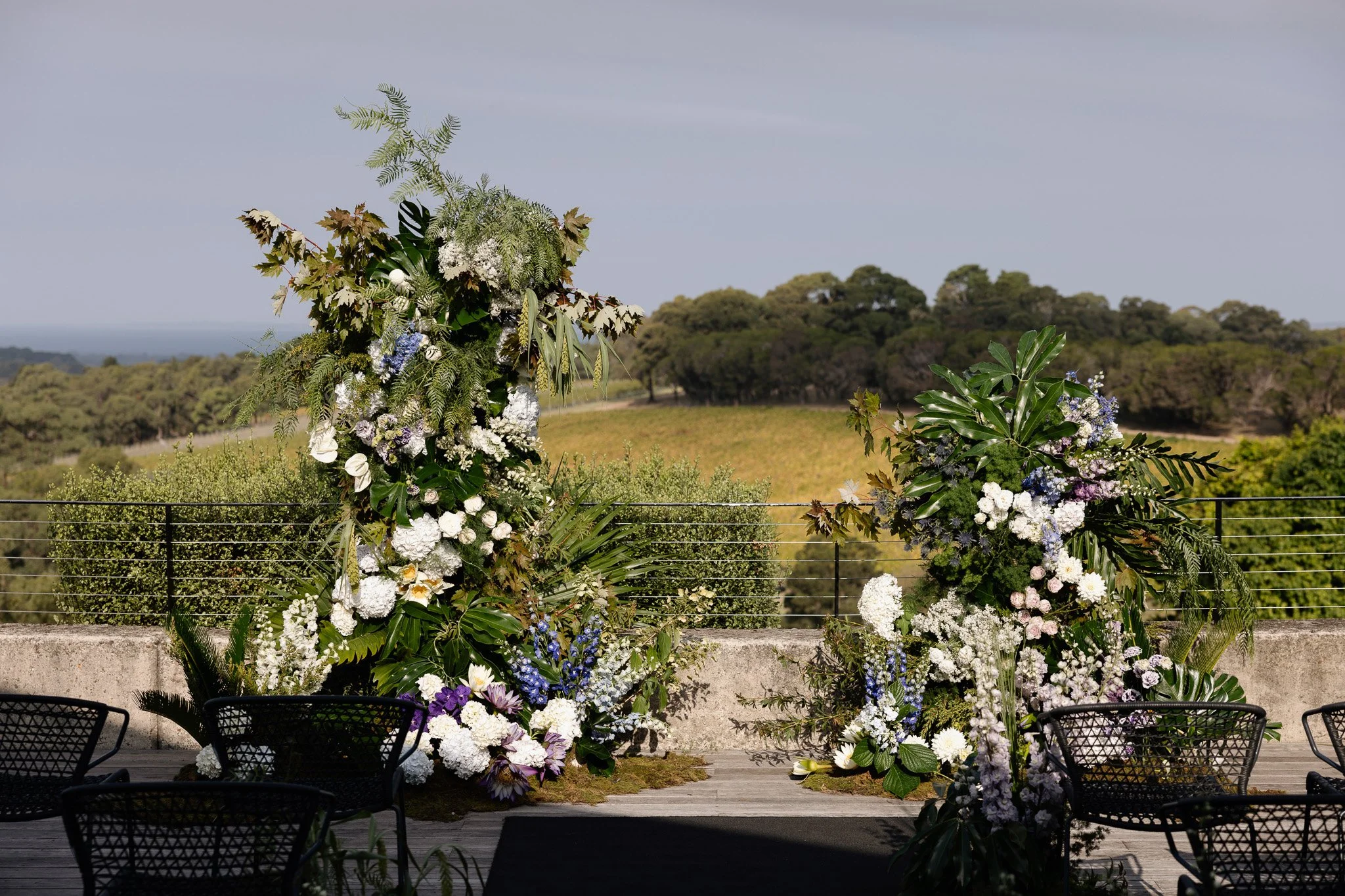 A floral arrangement for an outdoor event, featuring white, purple, and blue flowers with green leaves, set against a rural landscape with trees and a cloudy sky.