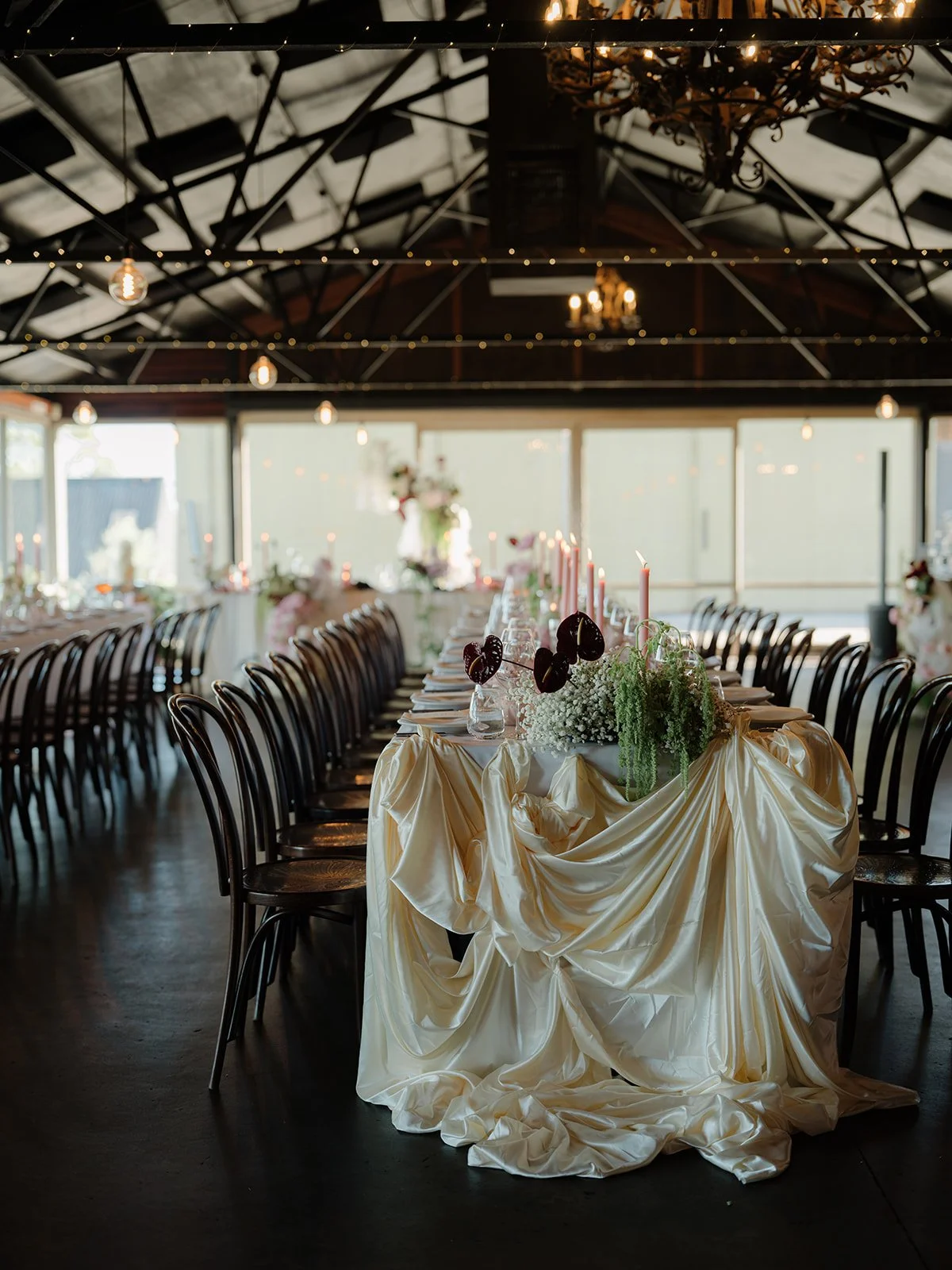 Elegant dining setup with a long table draped in a cream-colored satin cloth, decorated with pink candles, floral arrangements, and black heart-shaped decorations. The room features wooden chairs, string lights, and large windows, creating a warm, in
