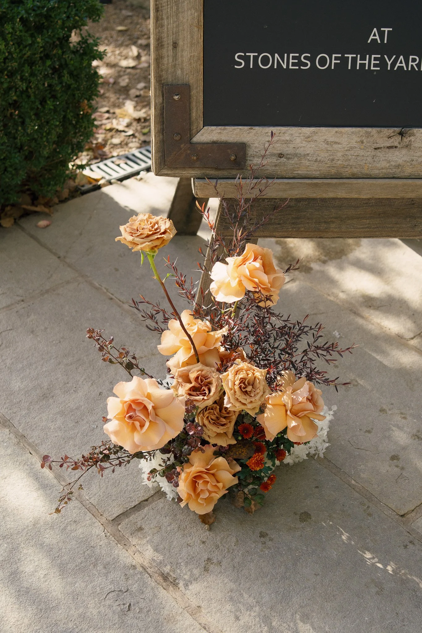A flower arrangement with peach-colored roses and other flowers and leaves placed on the ground next to a wooden sign with a blackboard surface, partially visible, with text reading 'At Stones of the Yard'.