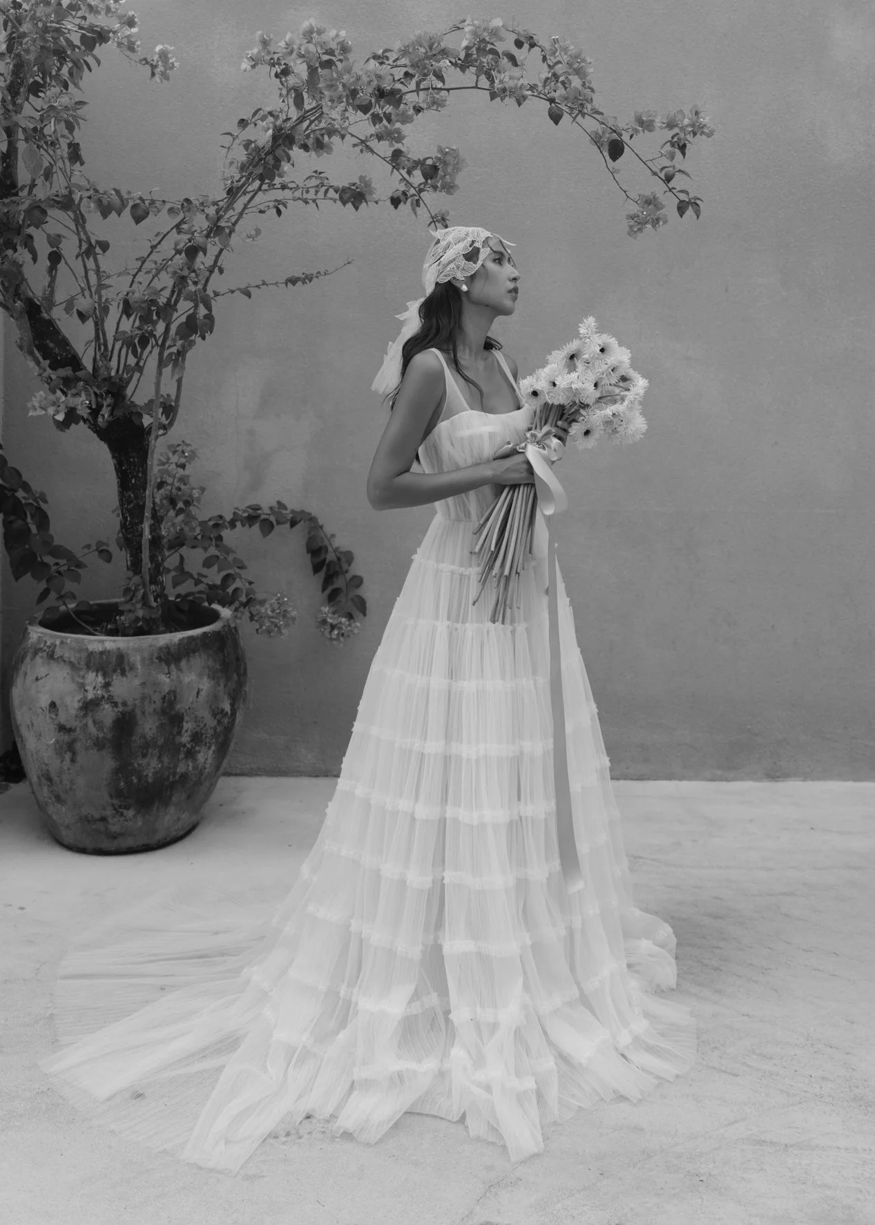 A woman standing in front of a plain background, holding a bouquet of flowers, dressed in a long, flowing white dress, wearing a head covering, and standing next to a large potted plant.