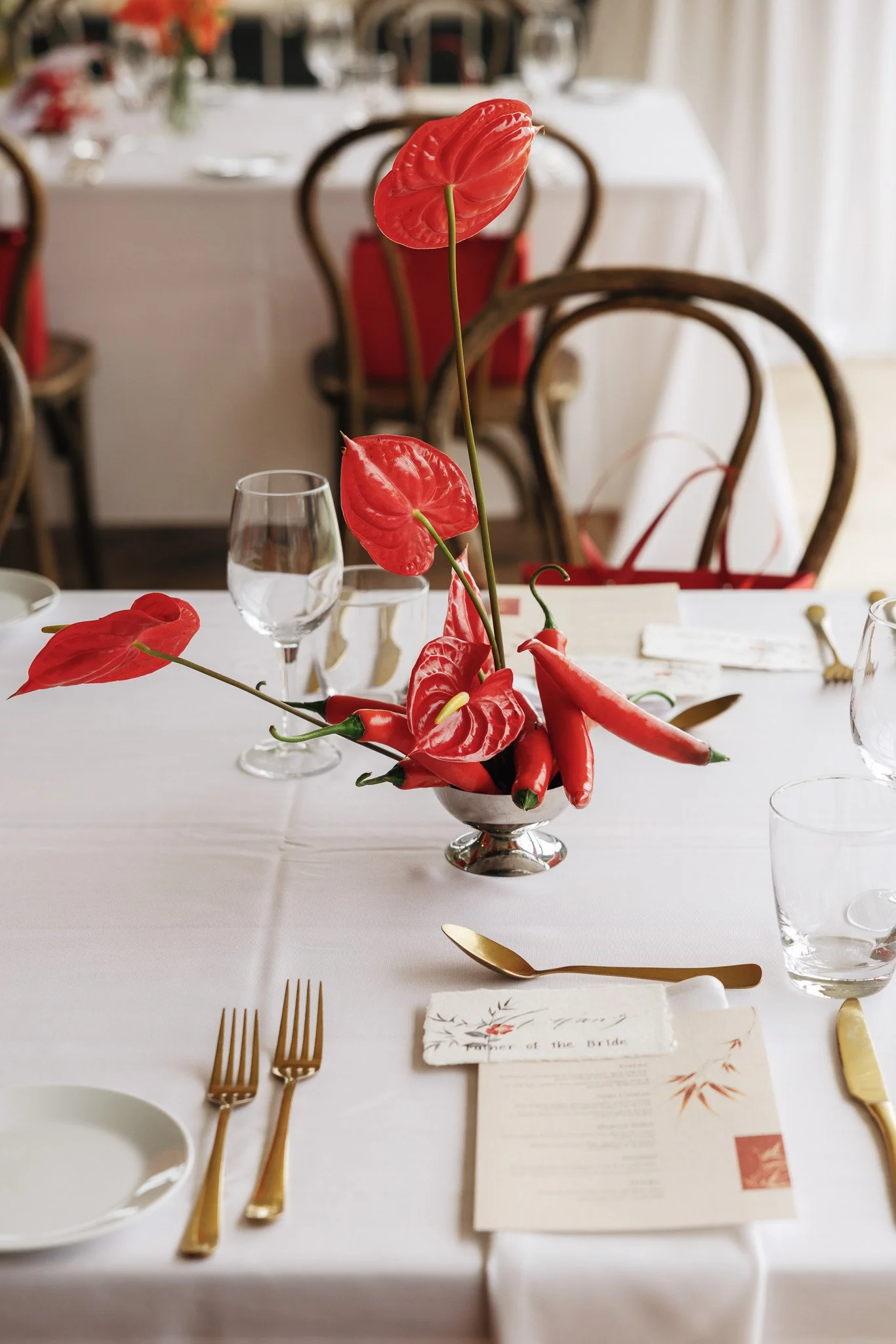 Formal wedding reception table setting with a centerpiece of red anthurium flowers in a silver bowl, gold flatware, a water glass, and printed menus.