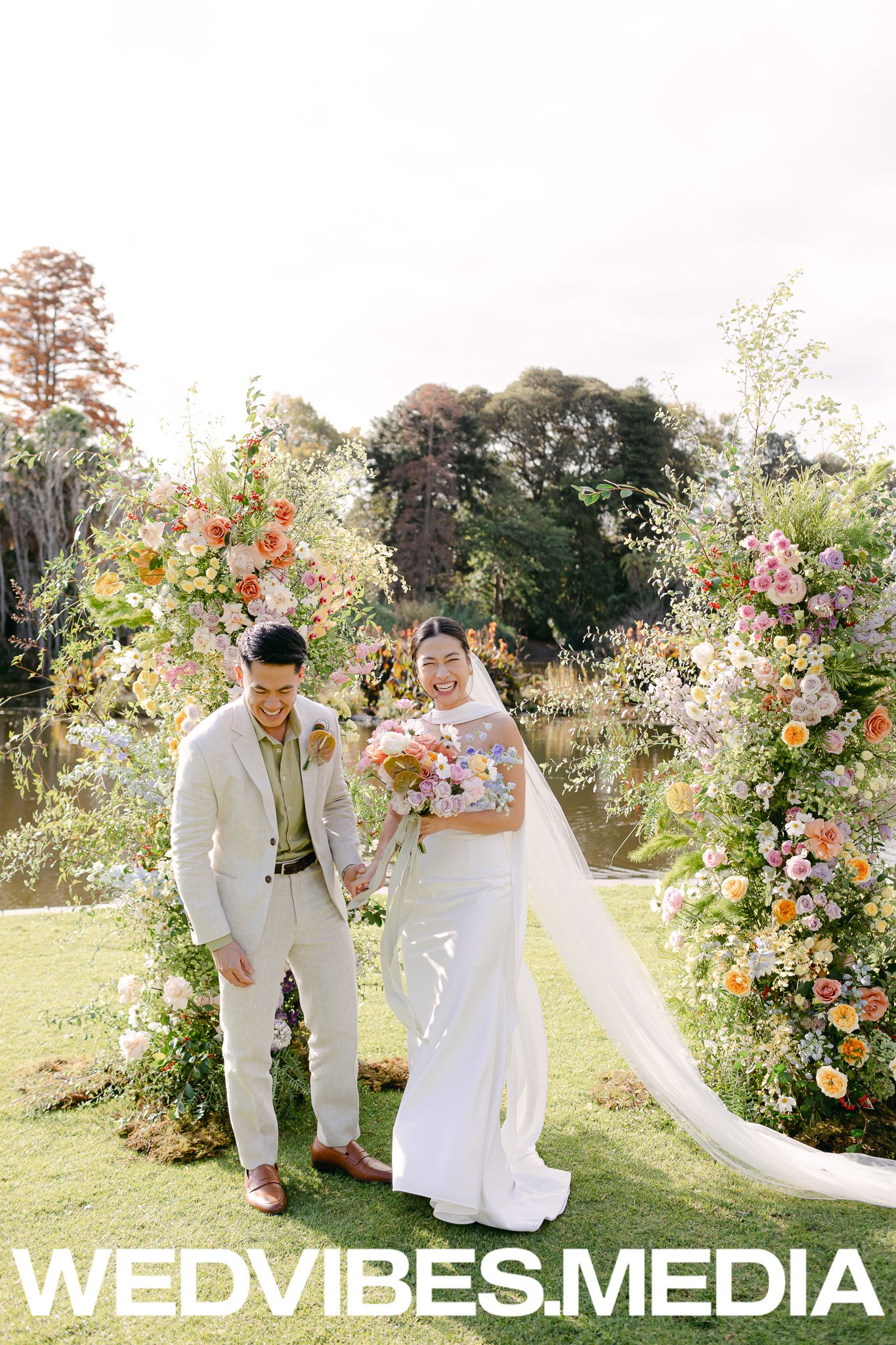 A happy bride and groom holding hands and smiling outdoors next to large floral arrangements during a wedding ceremony.