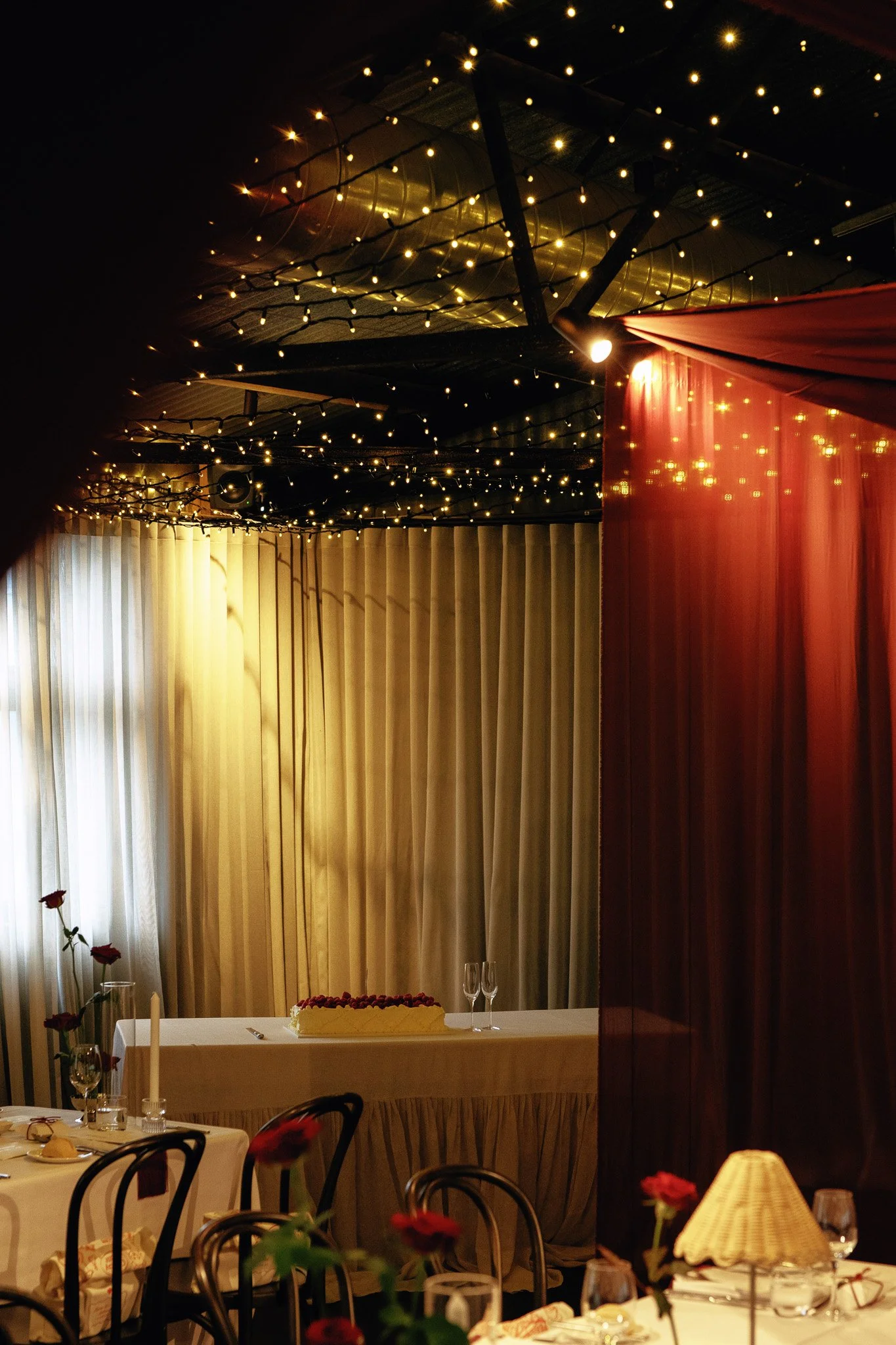 Indoor event space decorated with string lights on the ceiling, partially visible tables with wine glasses, candles, flowers, and a large white cake on a table.