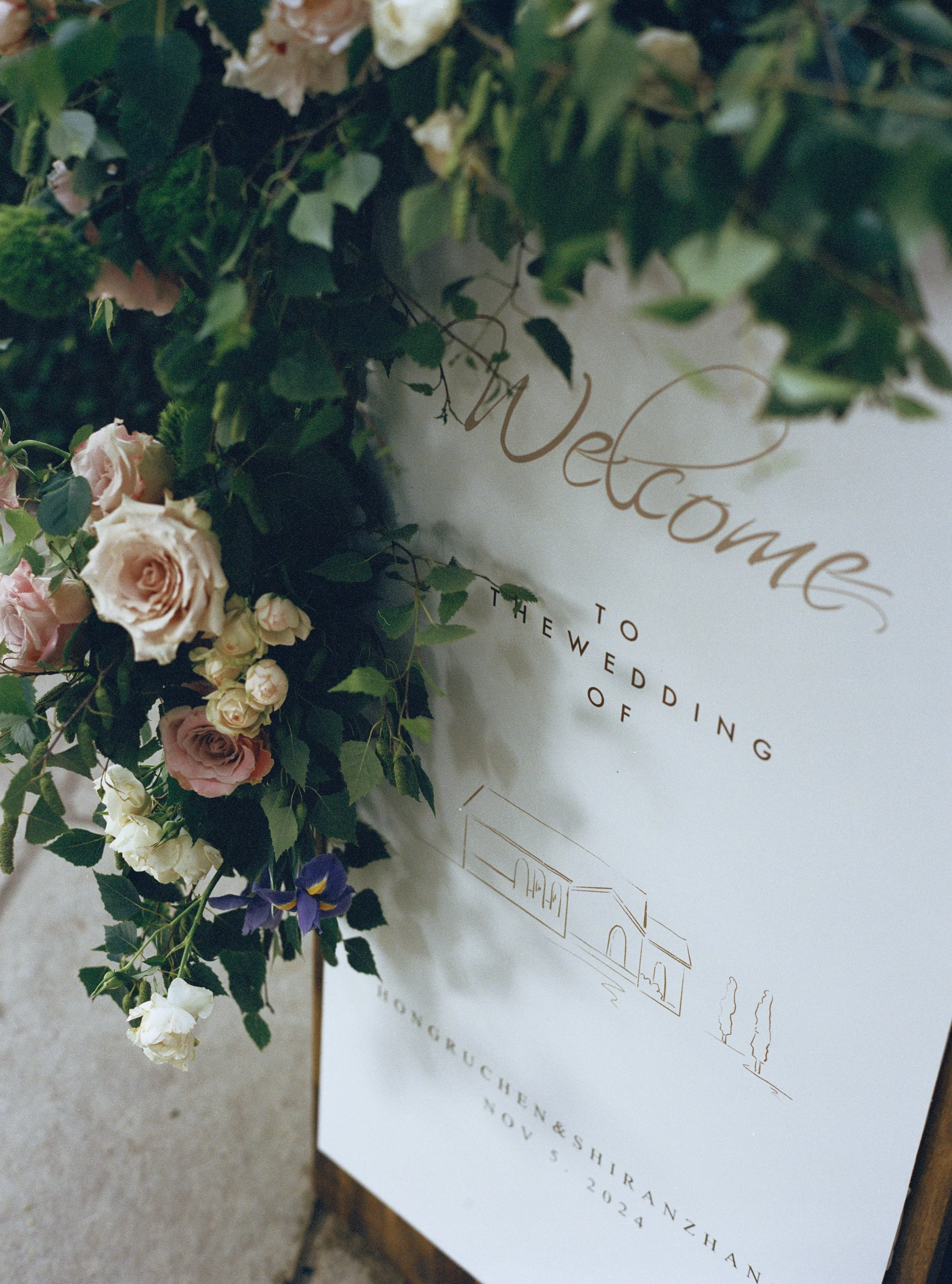 A floral arrangement with pink, white, and purple flowers and green leaves next to a white sign with gold lettering.