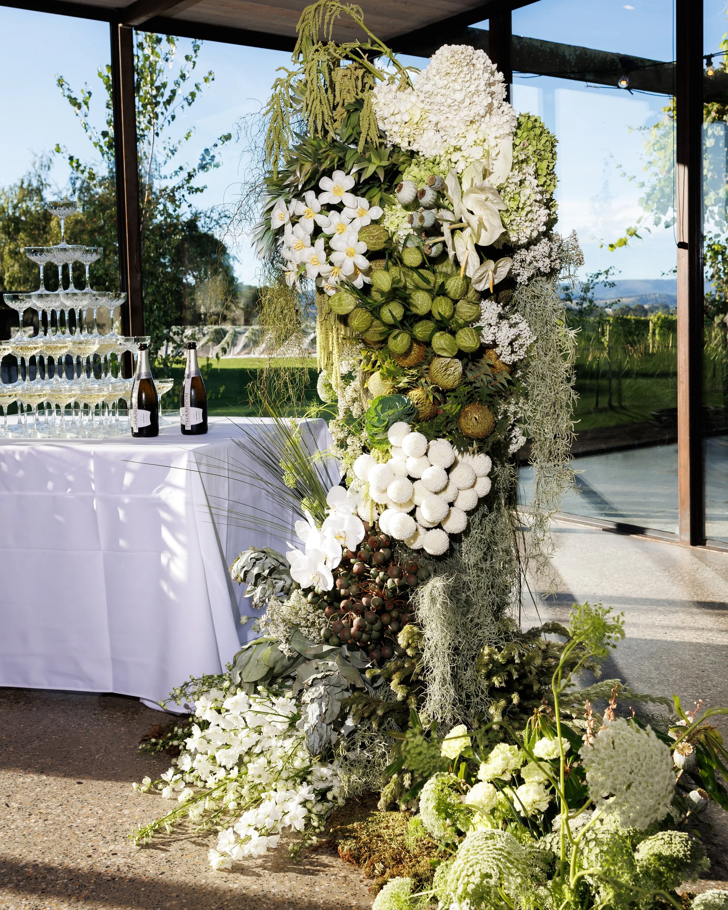 Tall floral arrangement of white and green flowers, including hydrangeas and orchids, set indoors with view of outdoor vineyards through large window.