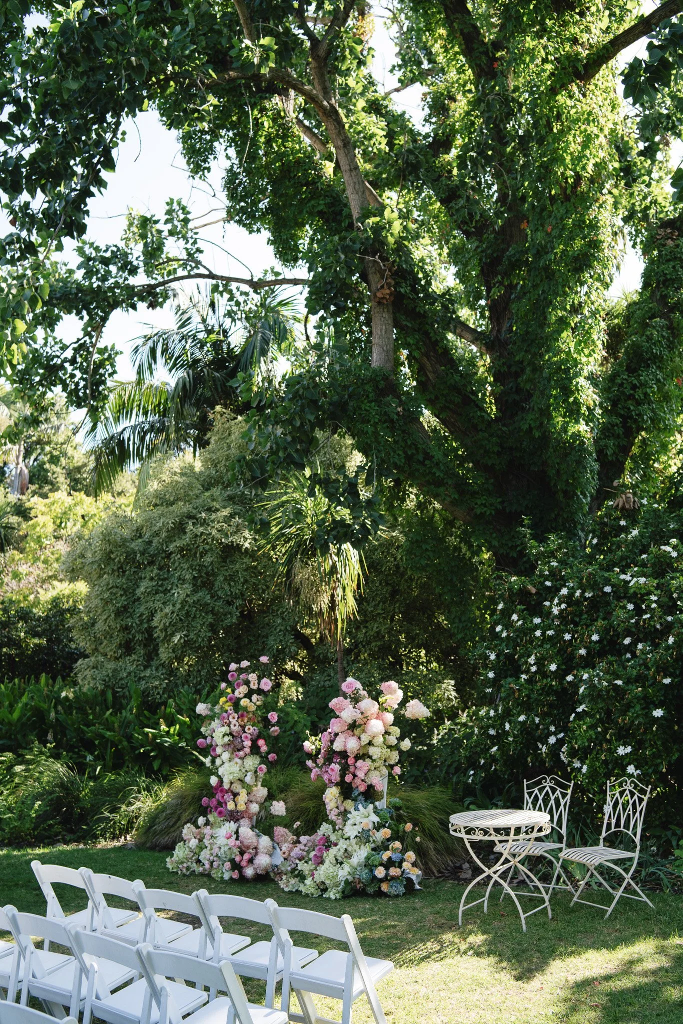 Outdoor garden wedding setup with white chairs arranged for guests, a floral arch with pink and white flowers, lush green trees and plants, and two white wrought iron tables and chairs on a sunny day.