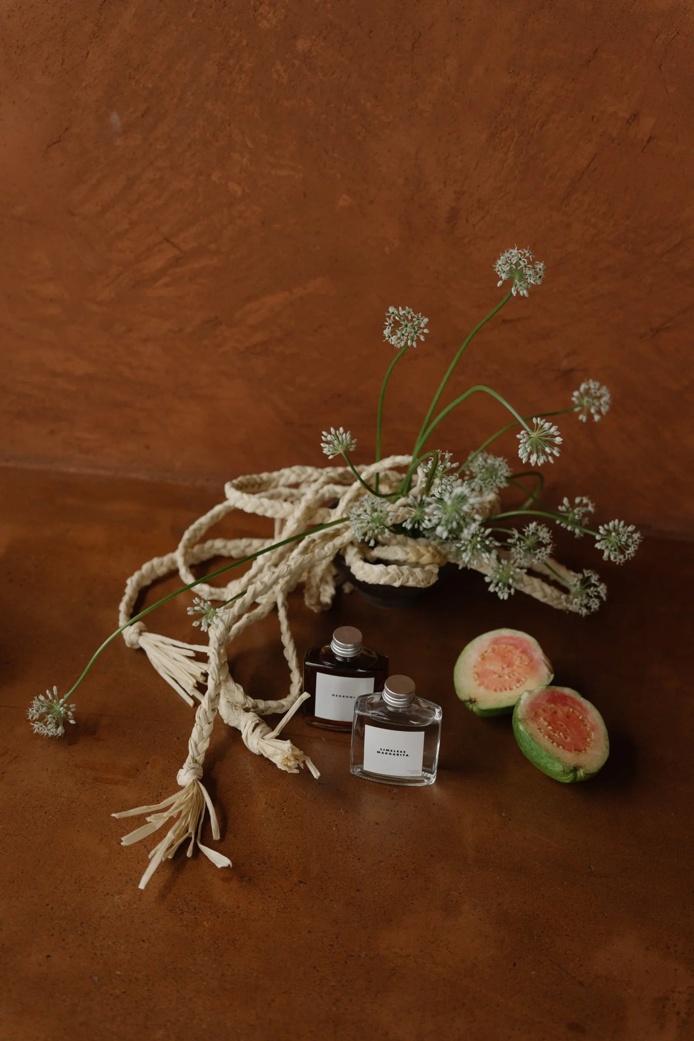 A small wicker basket with white floral accents, two perfume bottles, a halved watermelon showing pink flesh and seeds, and a few white flowers against a brown surface.