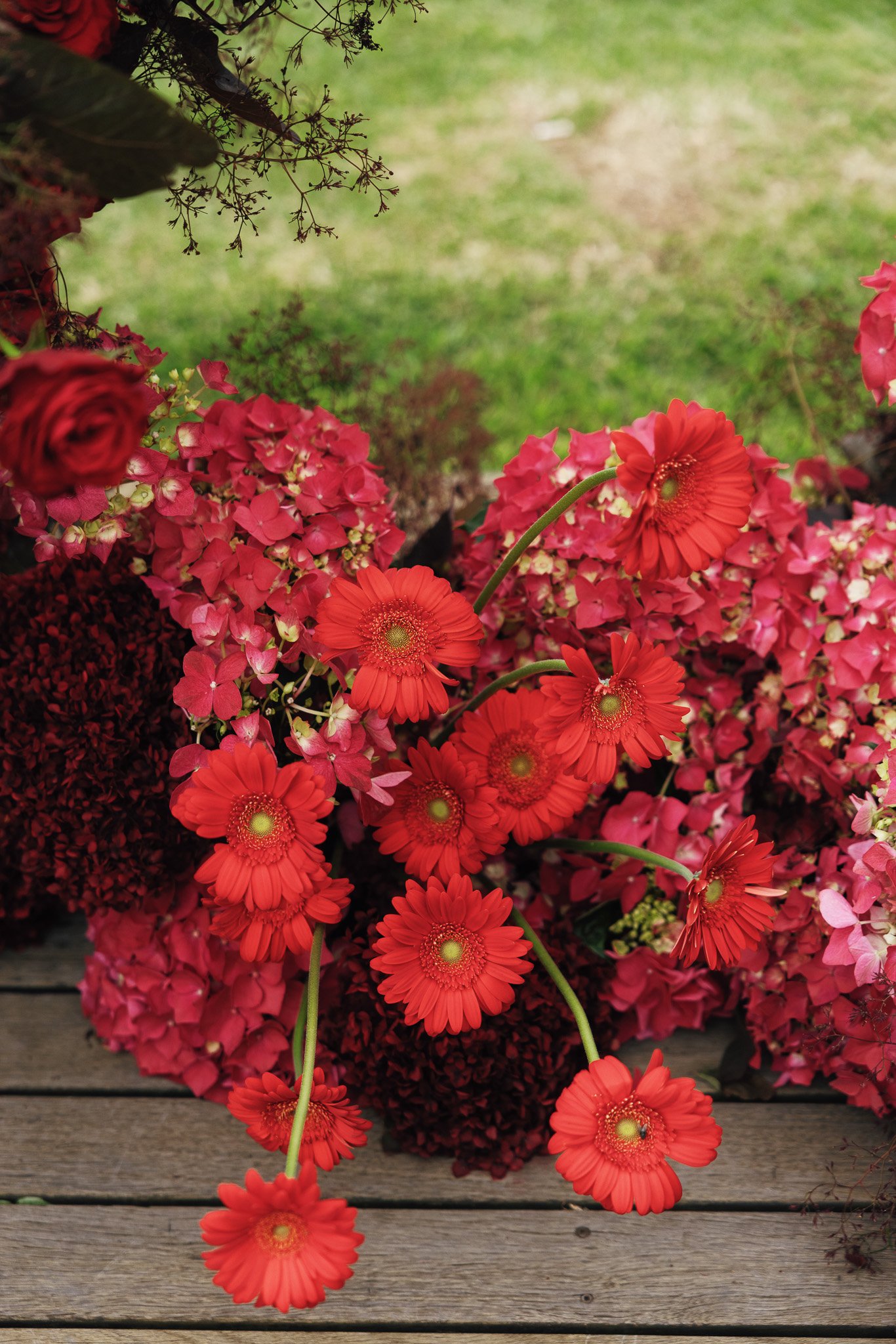 A bunch of vibrant orange and pink flowers sitting on a wooden surface with a blurred green grassy background.
