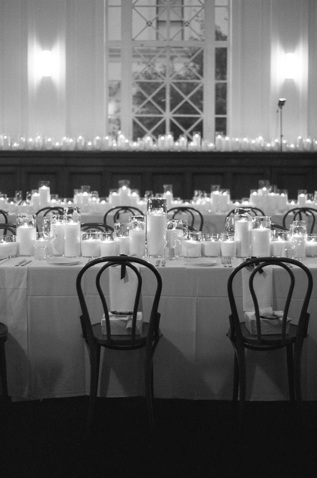 Elegant indoor event hall with long table decorated with candles and tableware, large windows with grid pattern, and chairs arranged for dining.