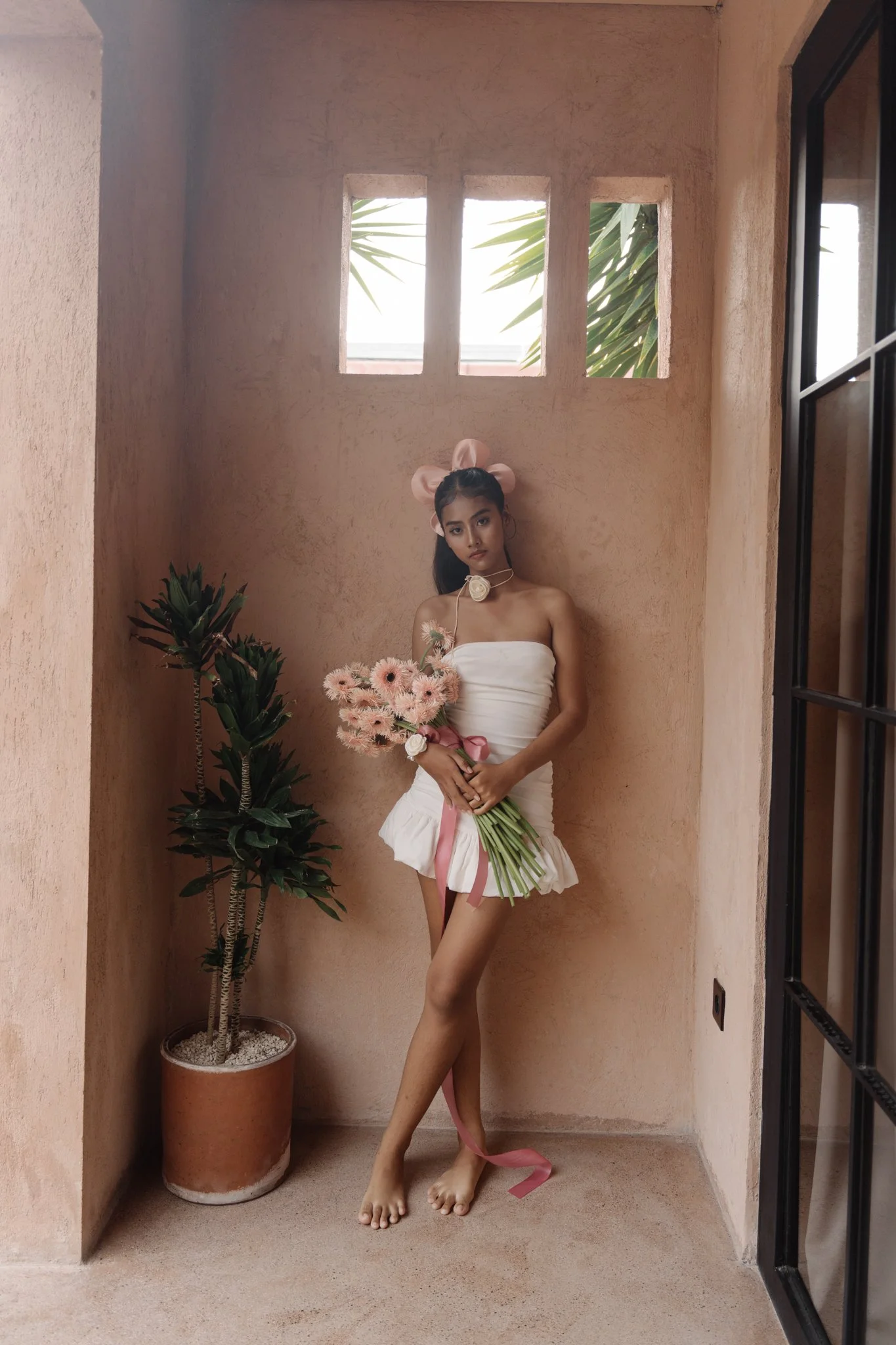 A woman with dark hair, wearing a strapless white dress, stands in a small, artistically lit room with peach-colored walls, holding a bouquet of pink flowers. She is barefoot, with one leg crossed in front of the other. She has a large pink bow headb
