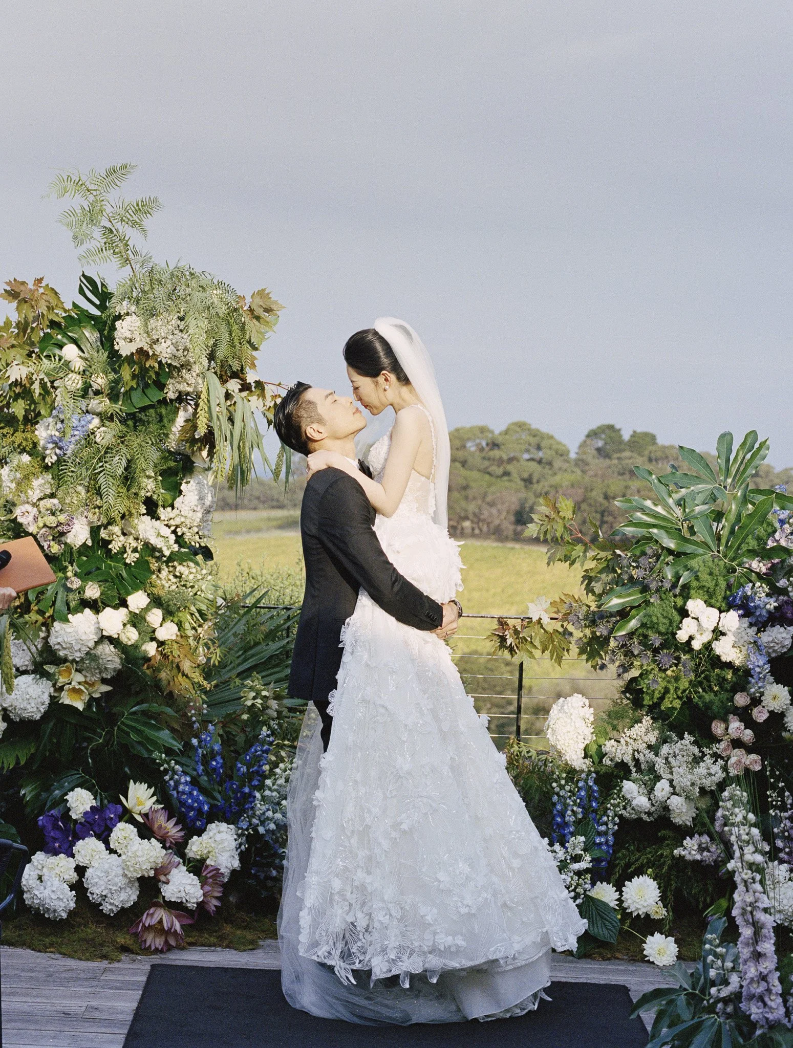 A bride and groom share a kiss at their outdoor wedding ceremony, surrounded by a floral arch and lush greenery in a scenic landscape.
