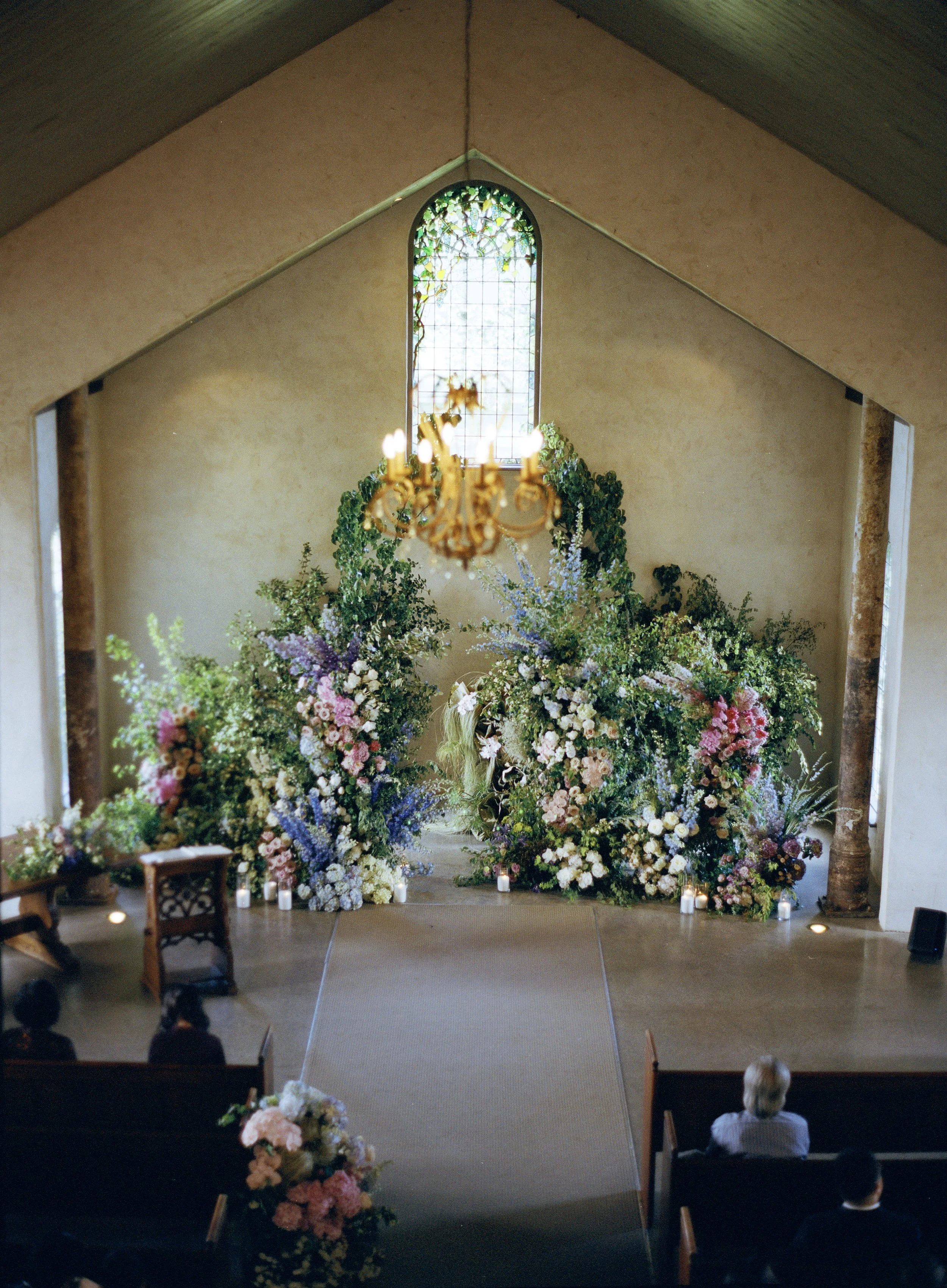 A decorated wedding altar inside a chapel with a large floral arrangement in front of a stained glass window, and a chandelier hanging from the ceiling above.