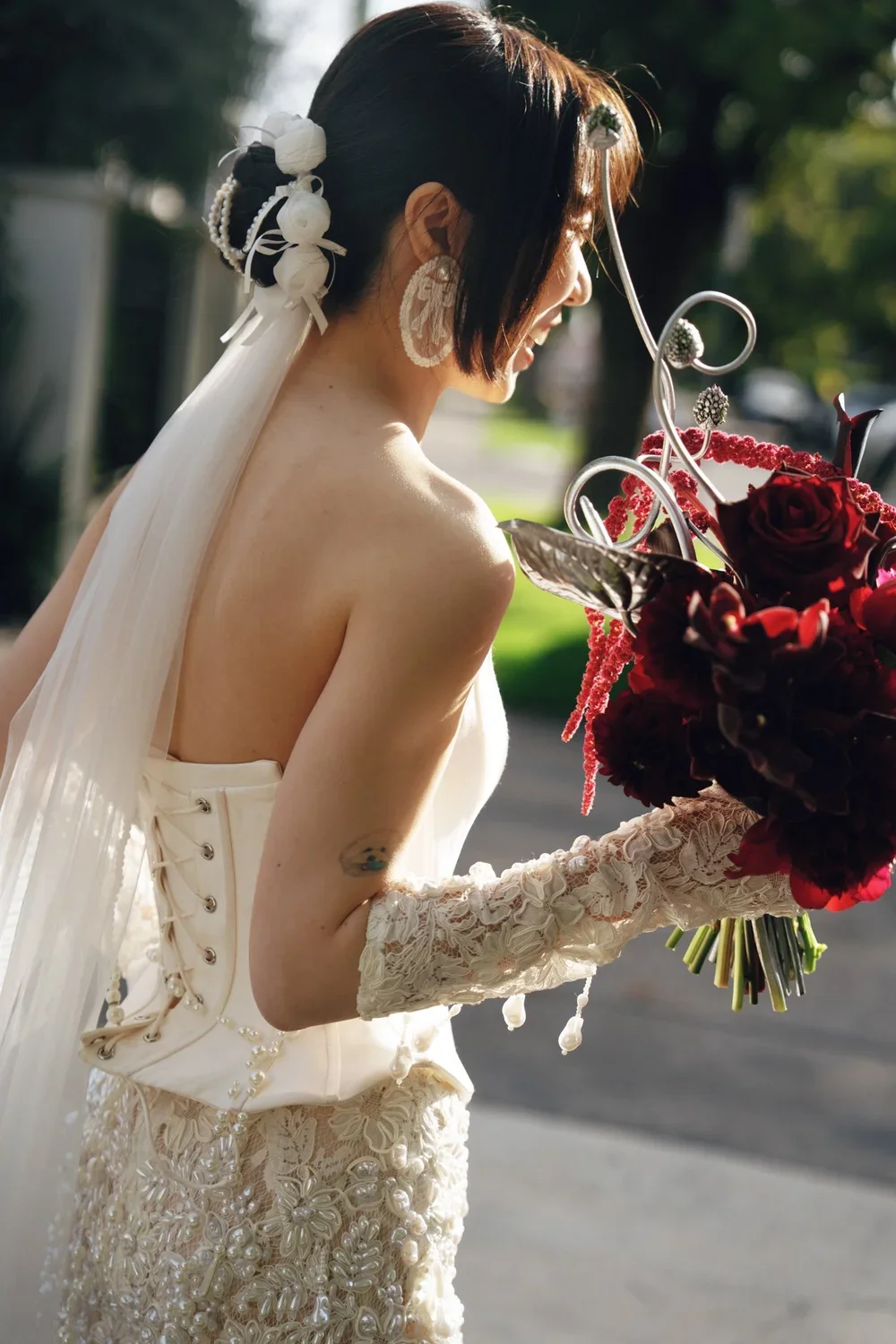 A bride with short dark hair, wearing lace gloves and a white dress, is holding a bouquet of dark red and pink flowers and smiling outdoors.