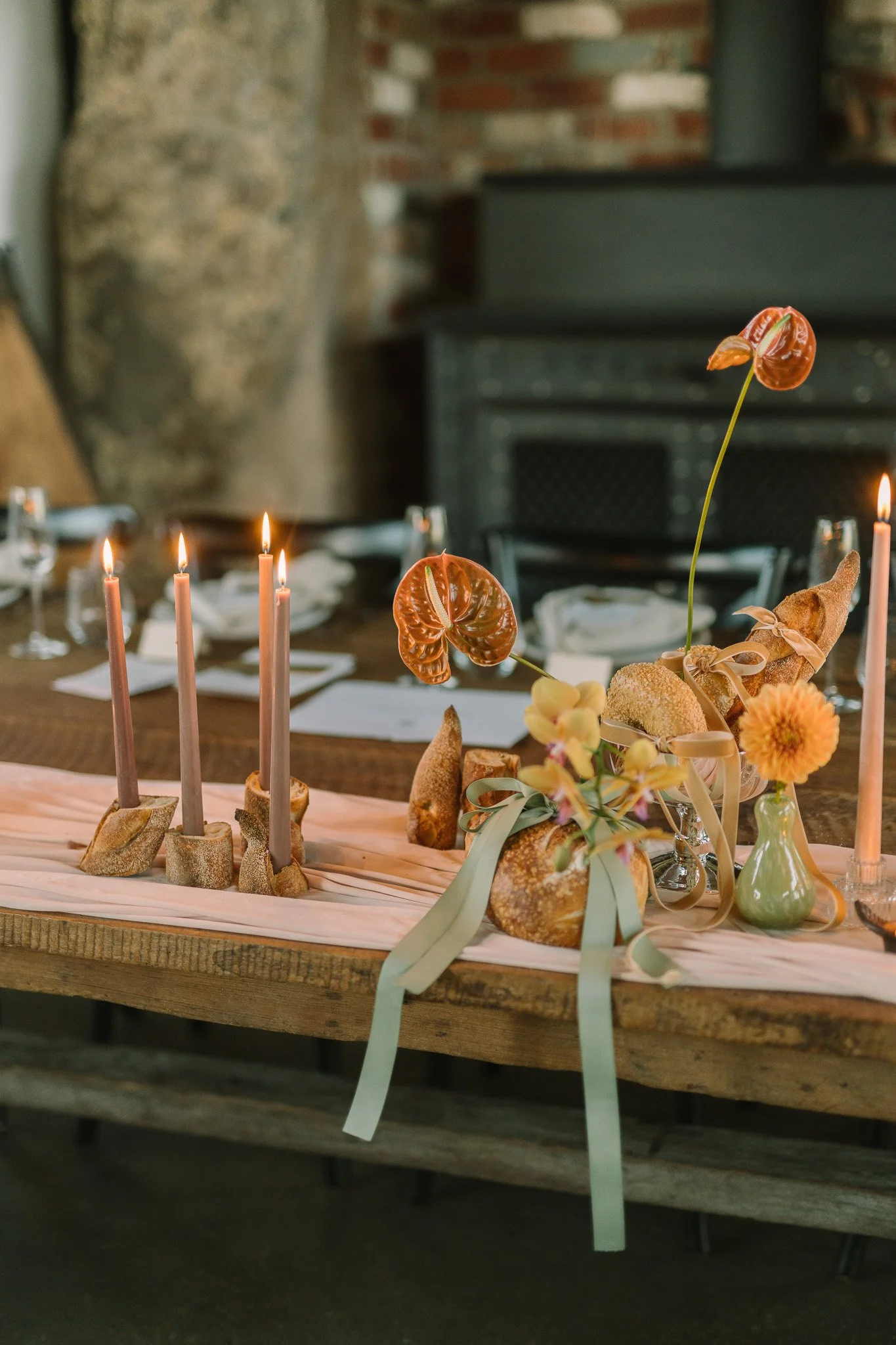 A table decorated with candles, bread, flowers, and pastries, set for a celebration or meal, with a blurred brick and stone background.
