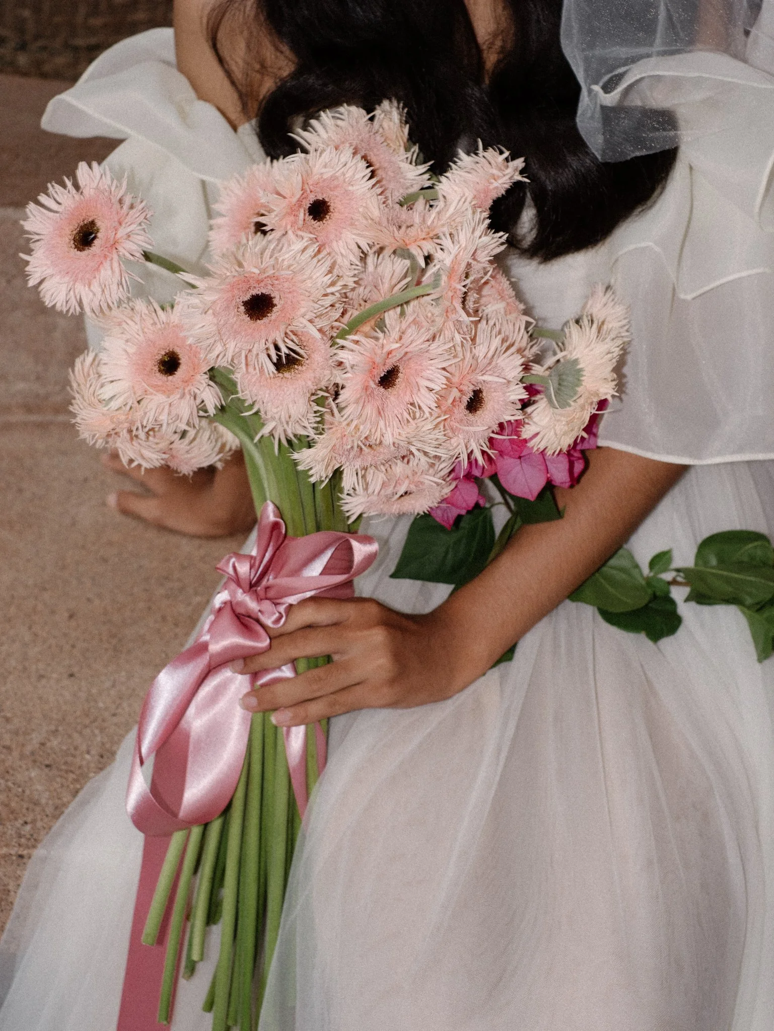 A girl in a white dress holding a bouquet of light pink and white flowers, sitting on a beige carpet. The bouquet is tied with a pink satin ribbon, and there are additional pink flowers visible.