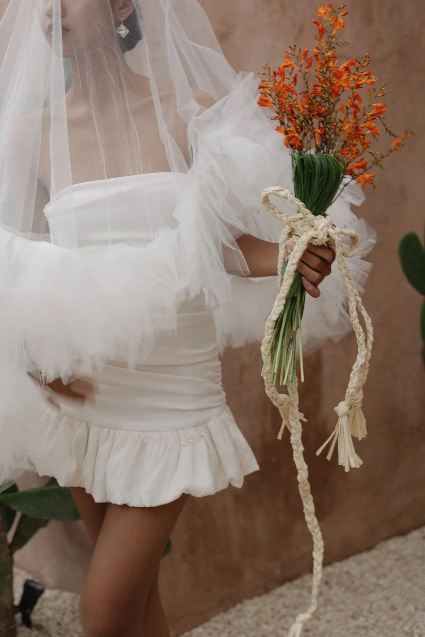 A girl dressed in a white dress with sheer fabric, holding a bouquet of orange flowers tied with a beige rope, in an indoor setting with a brown textured background.