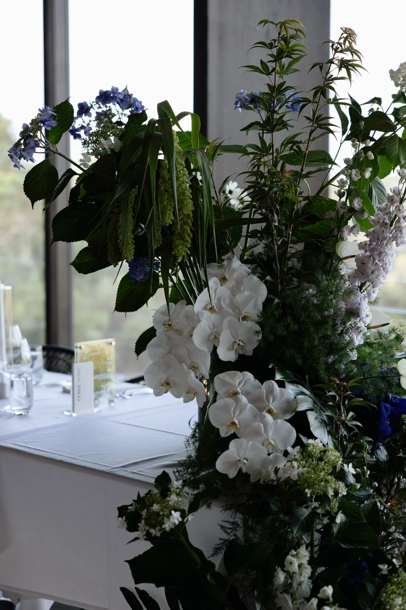 A floral arrangement featuring white orchids, blue hydrangeas, and various greenery on a table in front of a window with natural light.