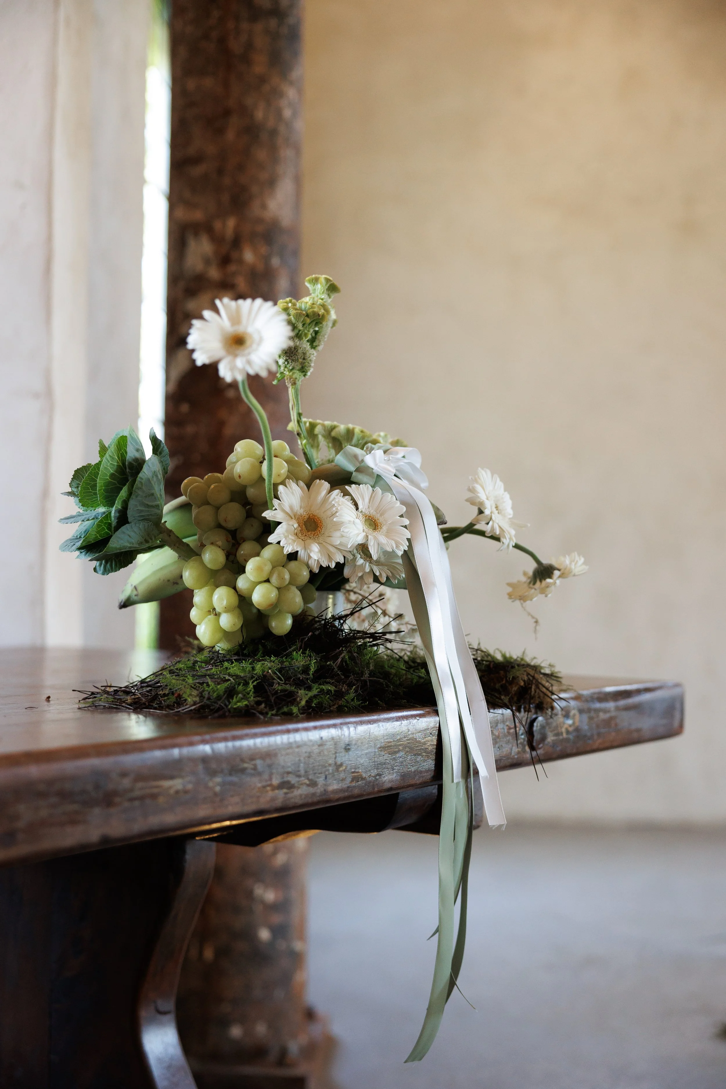 A floral arrangement with white daisies, greenery, and grapes, attached to a ribbon, placed on a rustic wooden table.