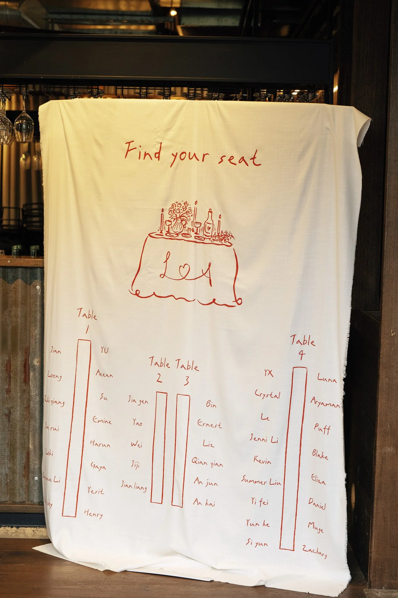 A large white cloth with red handwritten text and drawings. The writing says 'Find your seat' at the top and below it is an illustration of a table with a tablecloth, candles, wine glasses, and a flower arrangement. There are various handwritten name