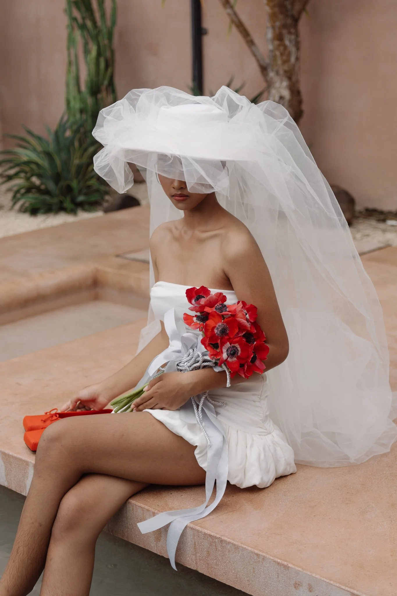 A woman wearing a large, white, tulle hat that covers her eyes, sitting on a beige stone bench in an outdoor setting with desert plants and rocks. She is dressed in a white outfit with a bouquet of red poppies, holding some orange objects, possibly s