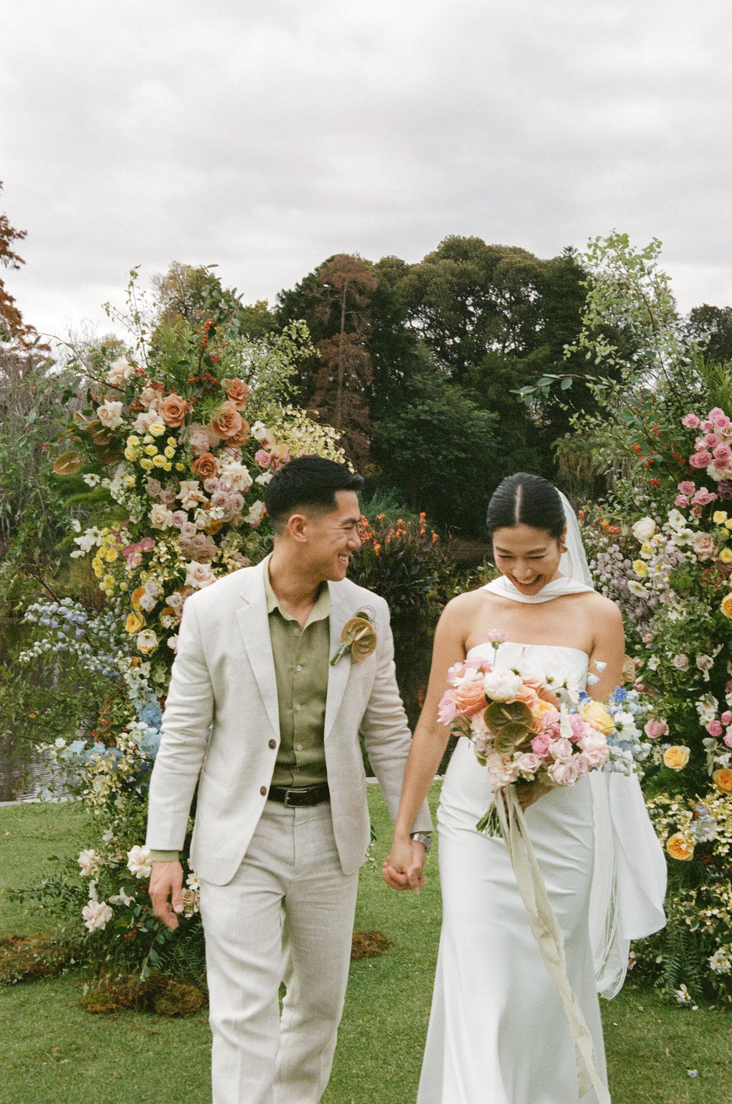 A bride and groom holding hands and smiling at each other during a wedding ceremony outdoors. The bride is wearing a strapless white wedding dress and holding a large bouquet of mixed pink, orange, and white flowers. The groom is dressed in a light-c