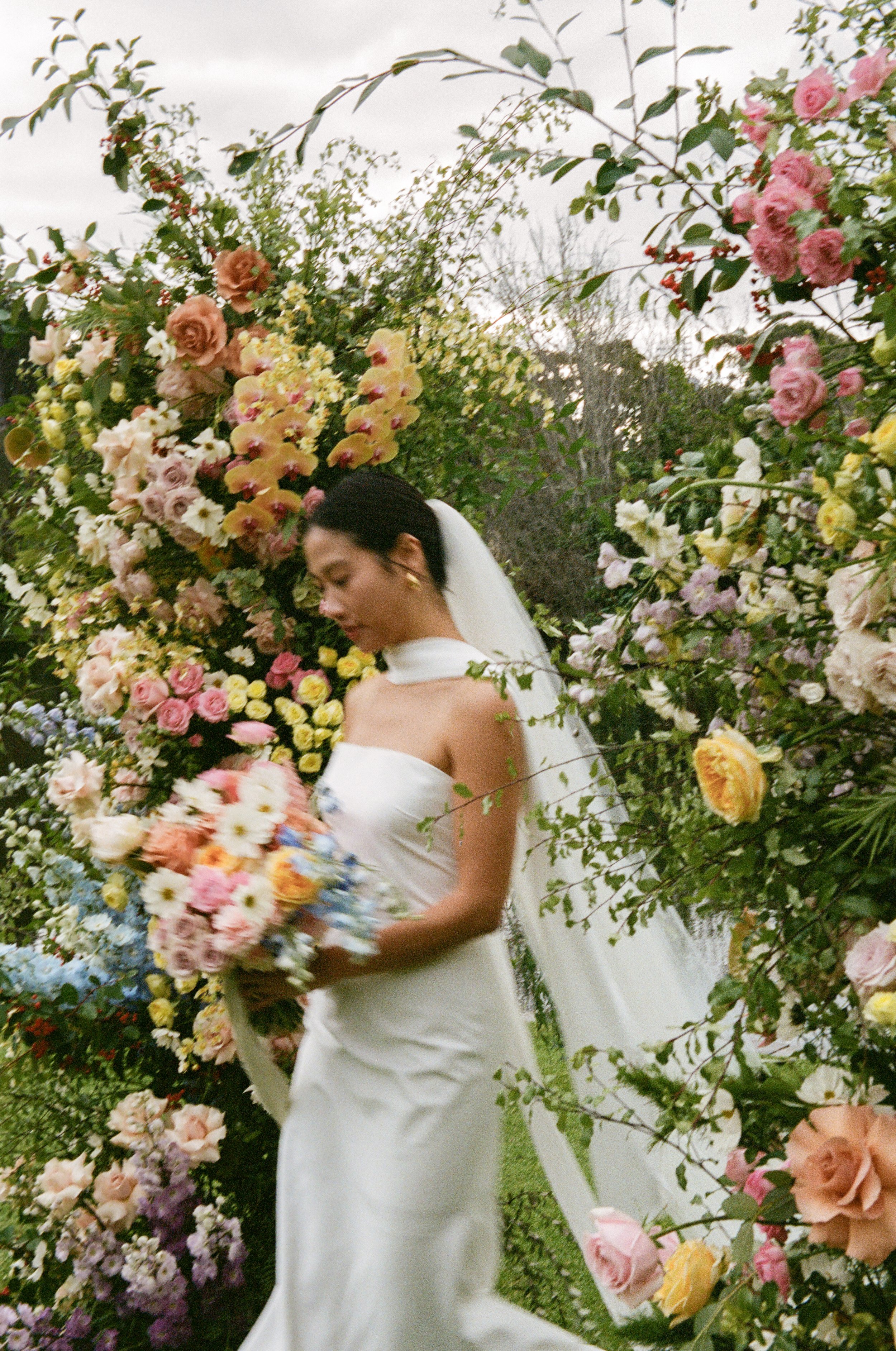 A woman dressed in a white wedding gown standing among a large floral arch made of pink, yellow, purple, white, and peach flowers.