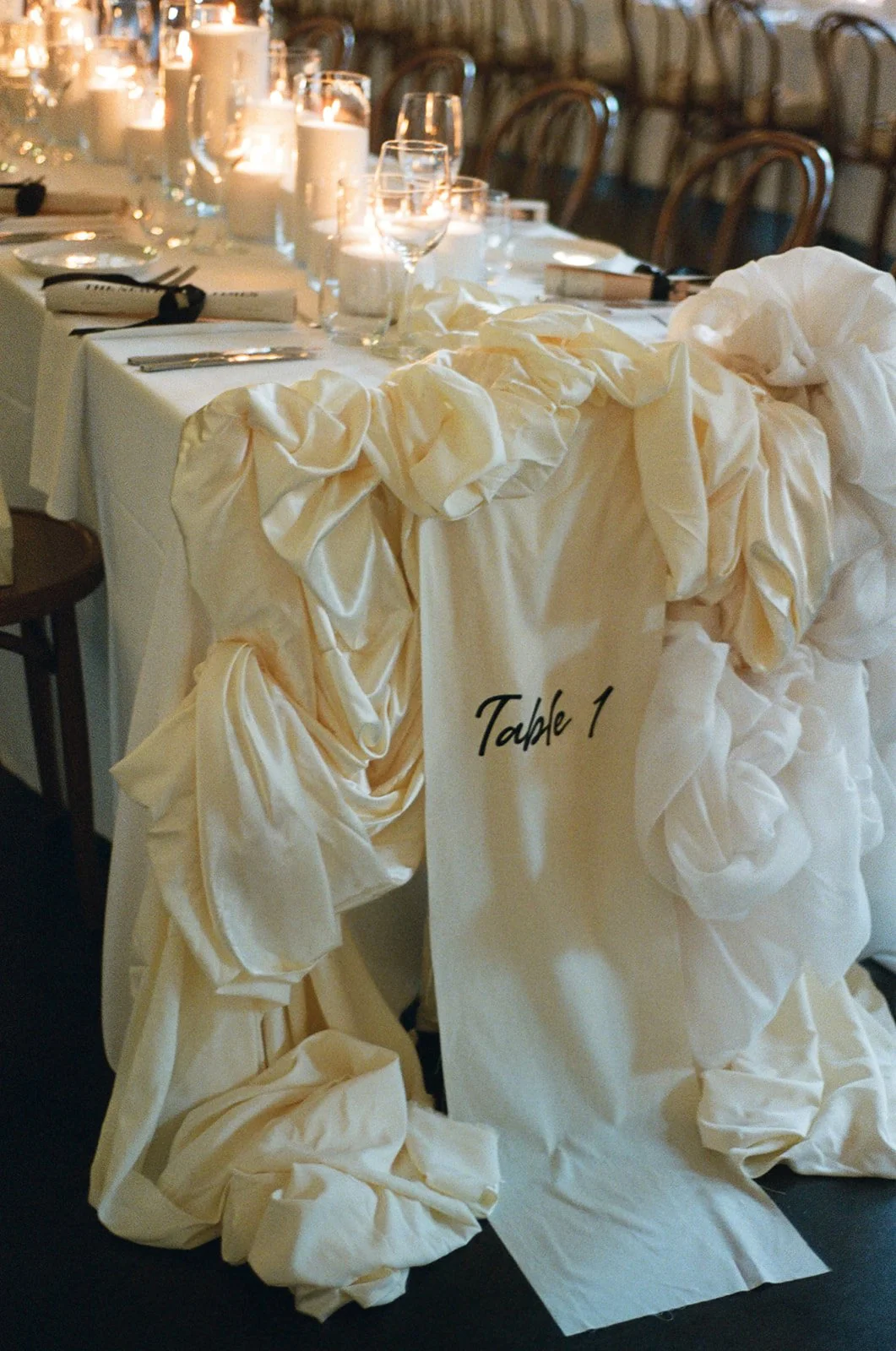 Wedding reception table decorated with candles and glassware. A chair draped with cream-colored fabric with a sign labeled 'Table 1' hangs on the back.