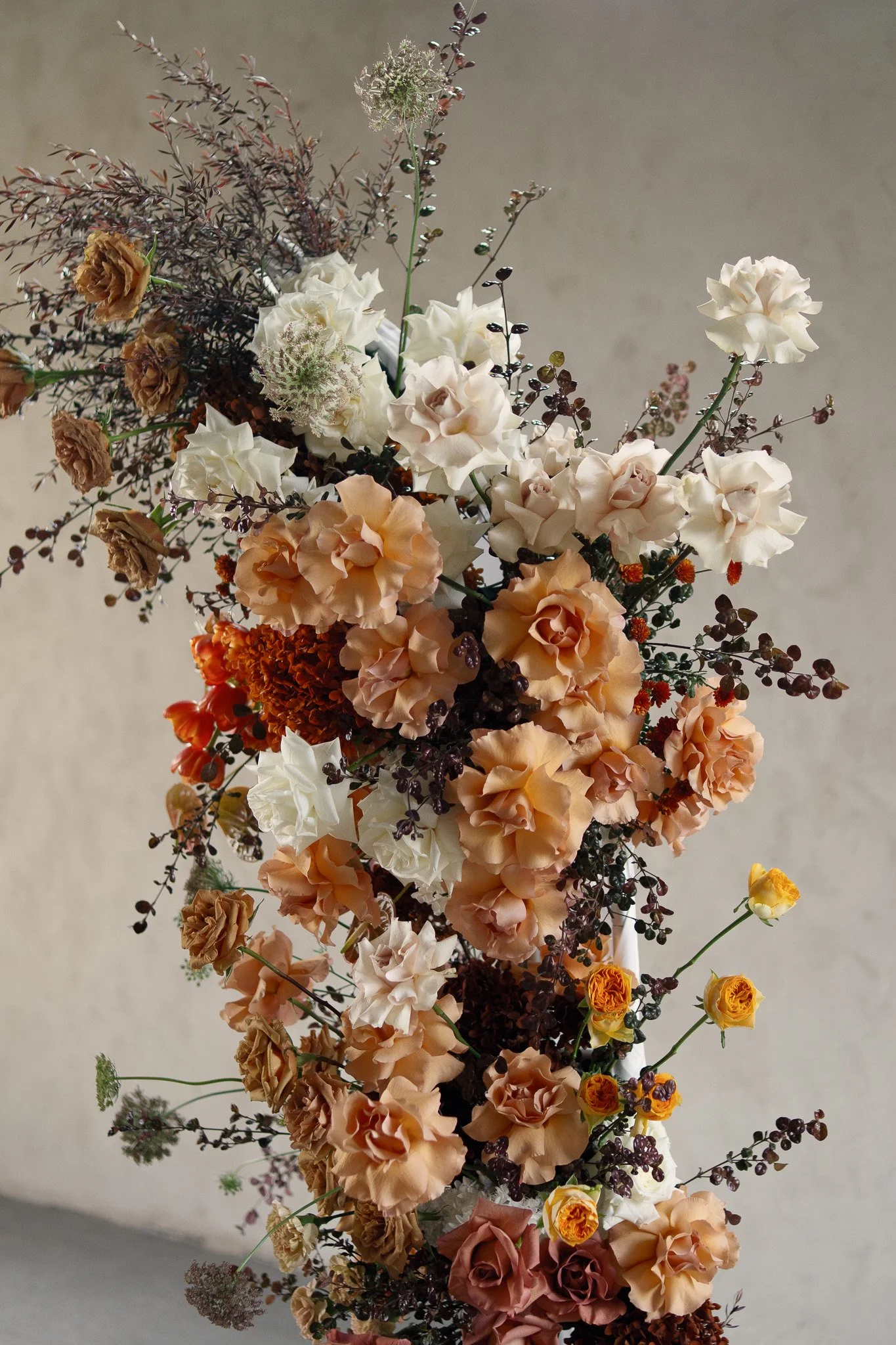 A large, colorful flower arrangement featuring cream, peach, and brown flowers with dark purple and green accents against a neutral background.