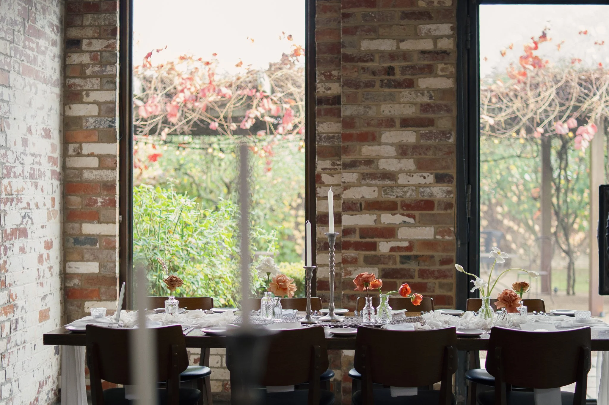 The interior of a room with exposed brick walls and large windows showing outdoor greenery and pink flowers. A table is set with flowers, candles, and tableware.