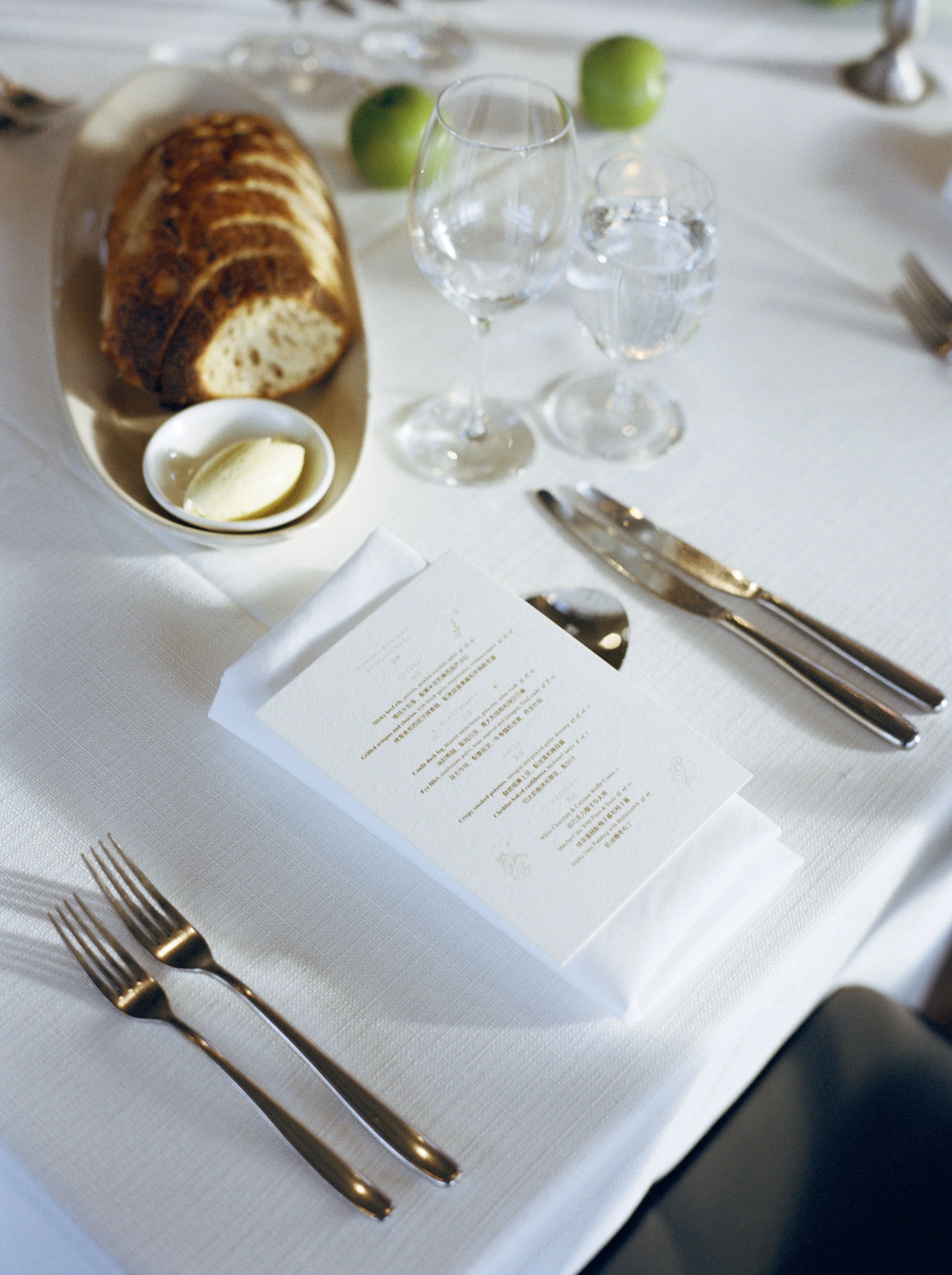 A table setting with two gold forks on a white tablecloth, a menu card, a white plate with a slice of bread, a small dish with butter, three glasses, and a menu or program paper.