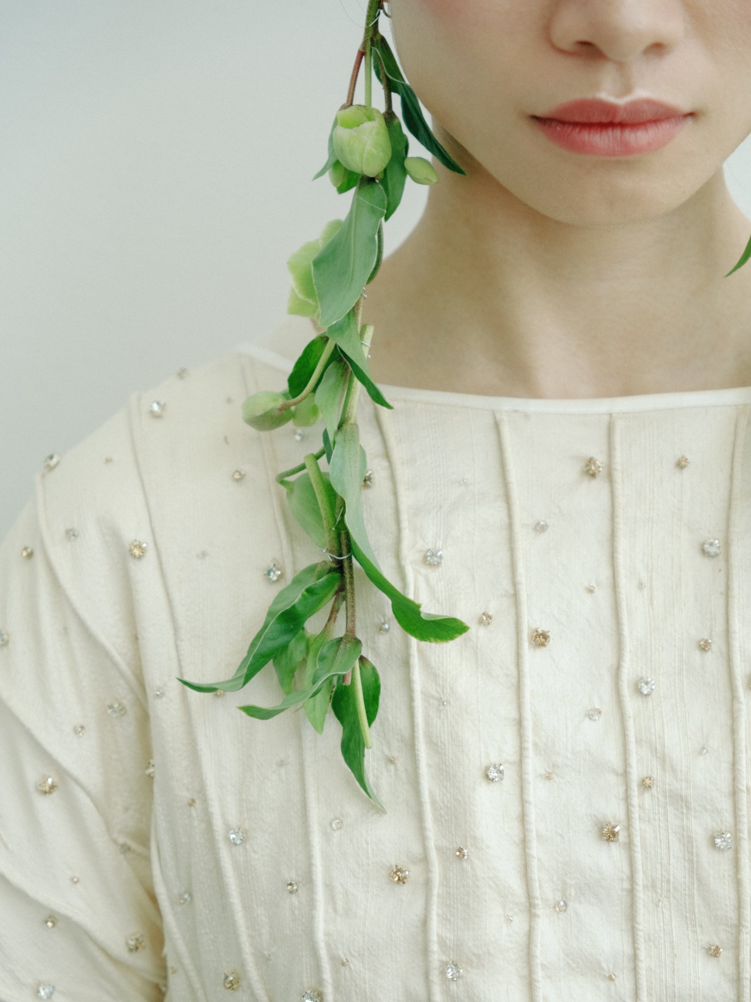 Close-up of a woman's face with a strand of green leaves and buds hanging near her cheek, dressed in a white dress decorated with small rhinestones.