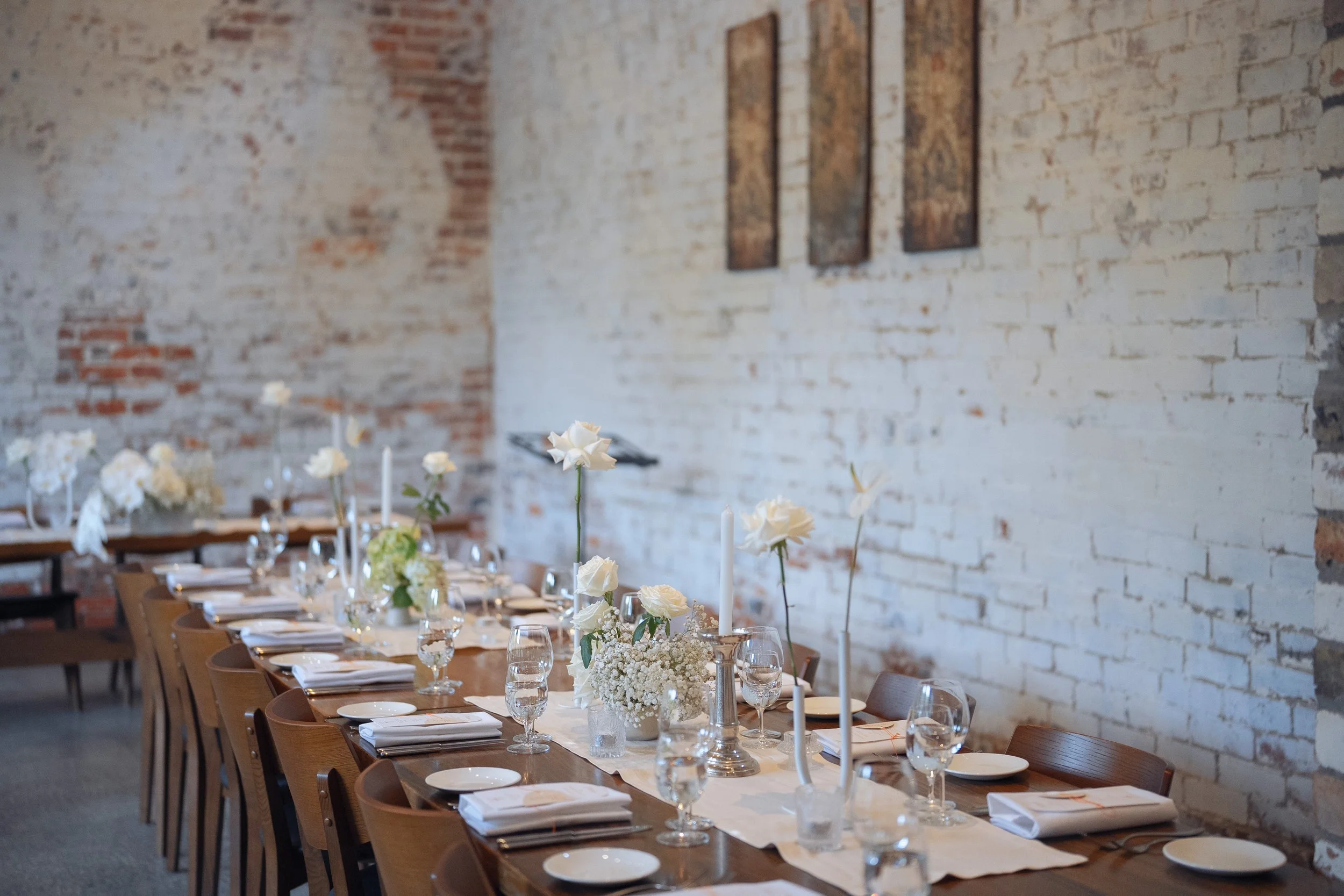 A long dining table set with white flowers in vases, candles, wine glasses, plates, and silverware, located against an exposed brick wall with three rectangular art pieces.