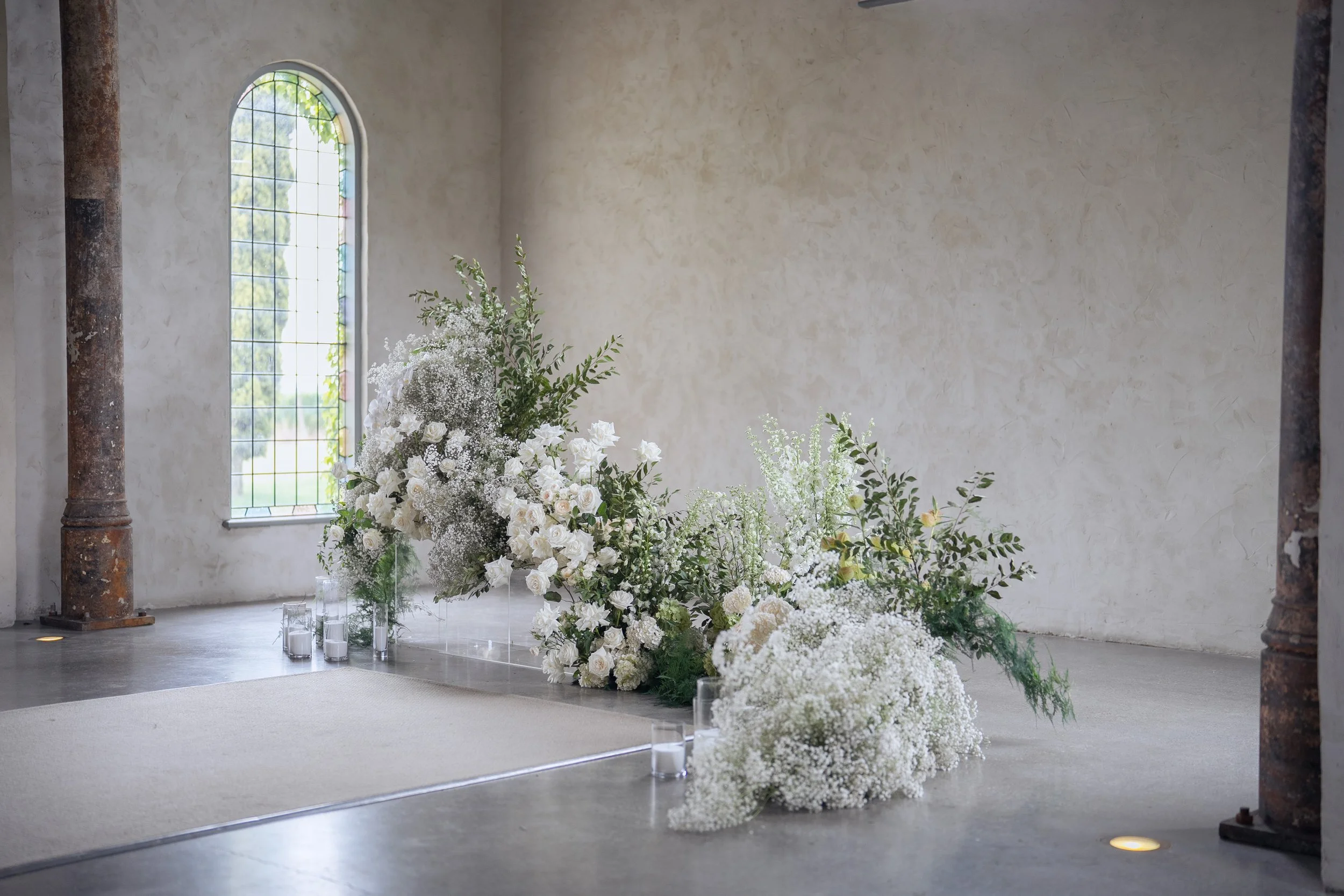 Elegant wedding or event floral arrangement with white roses, baby's breath, and greenery, placed on a platform inside a rustic building with tall arched windows and exposed rusted metal columns.