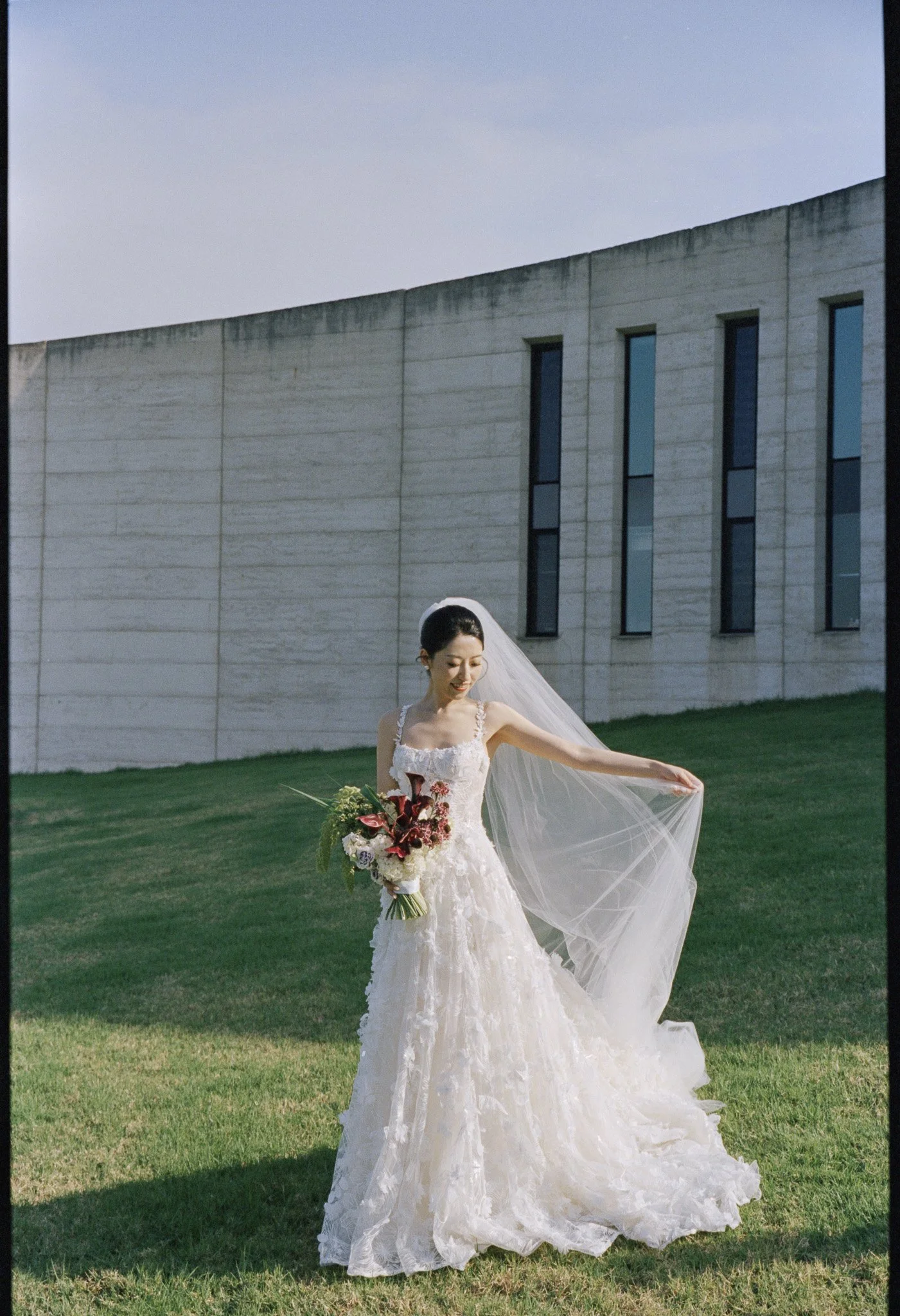 A bride in a white wedding gown holding a bouquet of flowers stands on a grassy lawn outside a modern building with tall narrow windows, a sloped roof, and gray stone walls, under a cloudy sky.