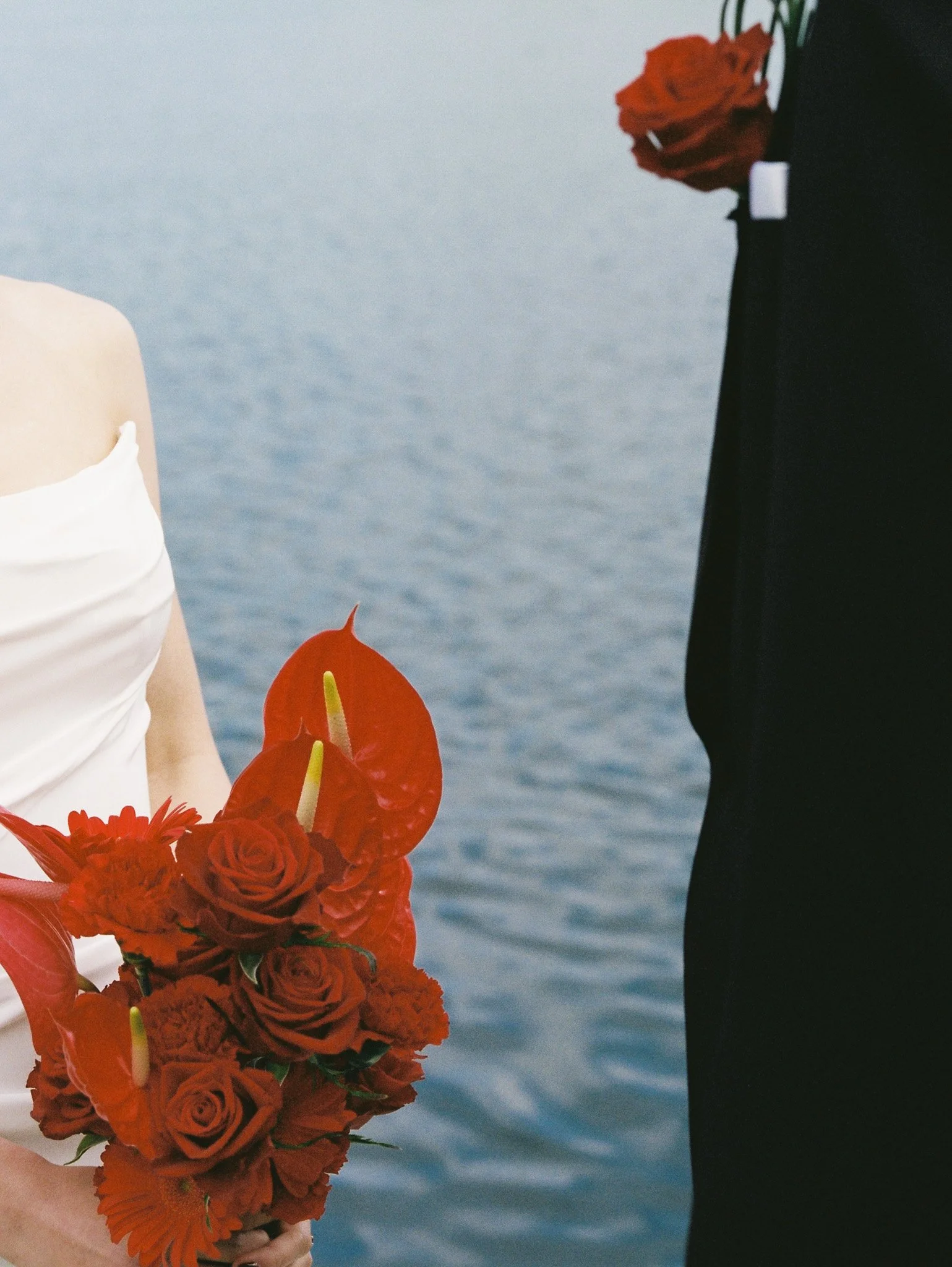 A bride and groom are holding red floral bouquets by the water, with the bride on the left and the groom on the right, holding hands.
