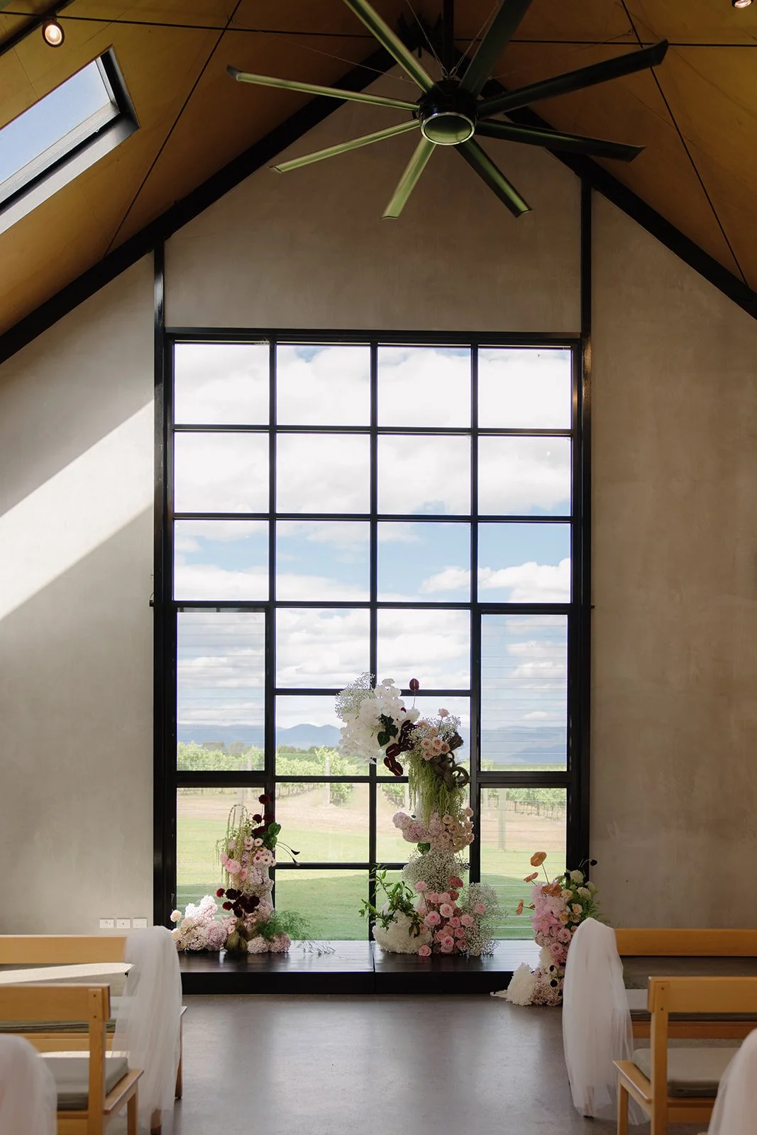 A large interior space with a tall window overlooking a rural landscape, decorated with floral arrangements for a wedding or event.